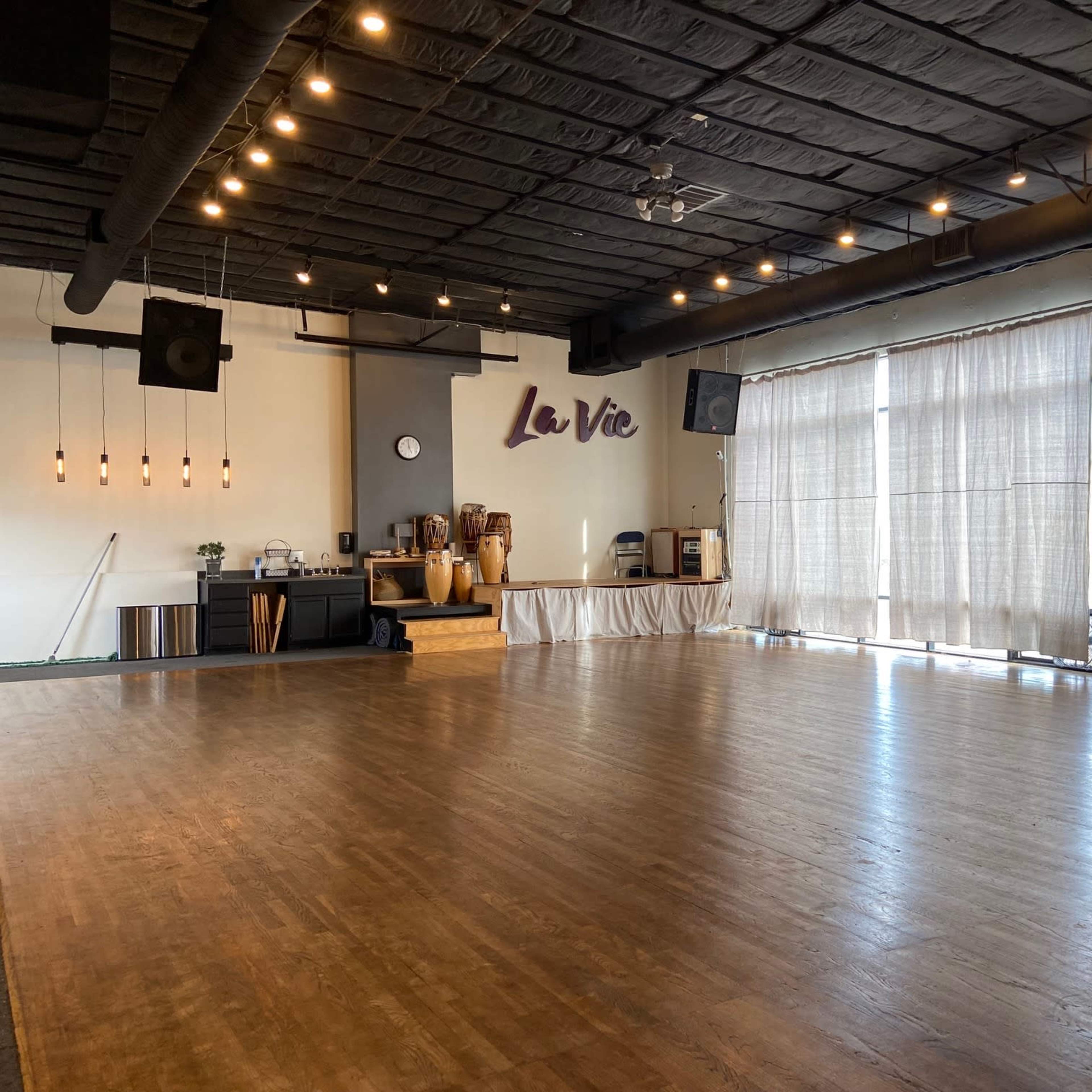 An empty dance studio with wooden flooring, large windows covered by sheer curtains, and a wall featuring the name "La Vie."