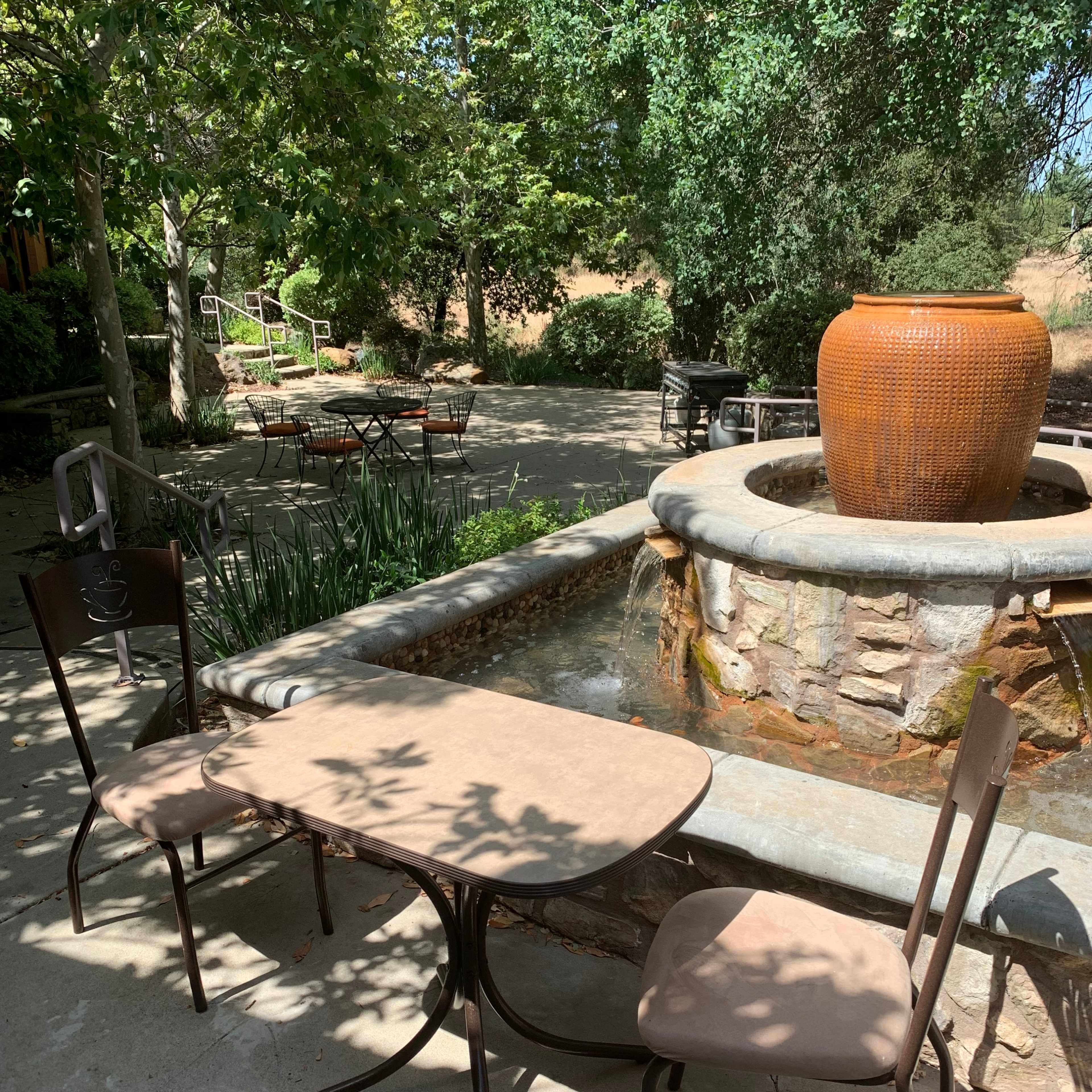 A small outdoor seating area features two chairs and a table beside a stone fountain surrounded by greenery.