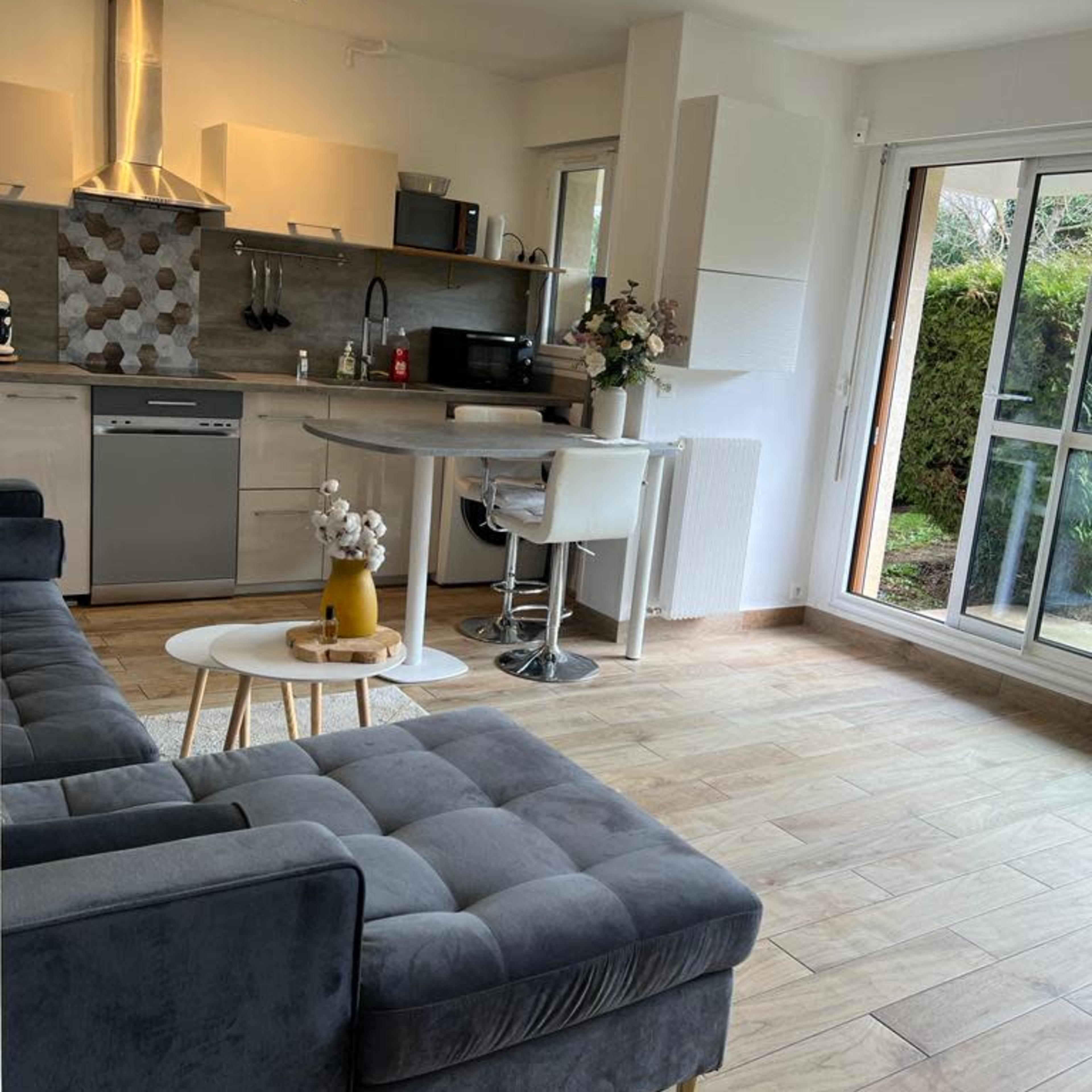 A modern kitchen and living area with a gray sectional sofa, a white coffee table, and a view of a garden through large glass doors.