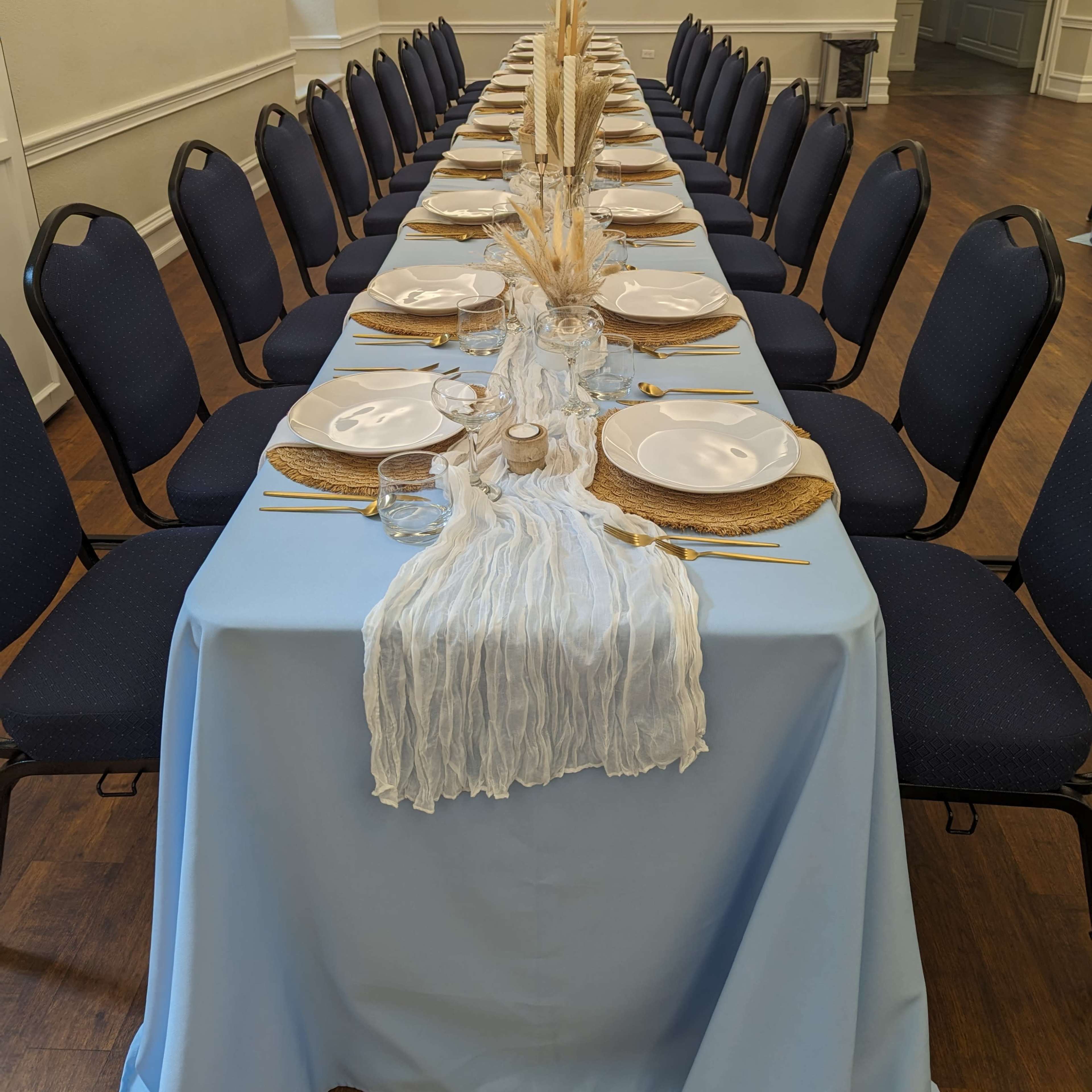 A long dining table is set with blue tablecloths and arranged for a meal, featuring plates, utensils, and decorative elements down the center.