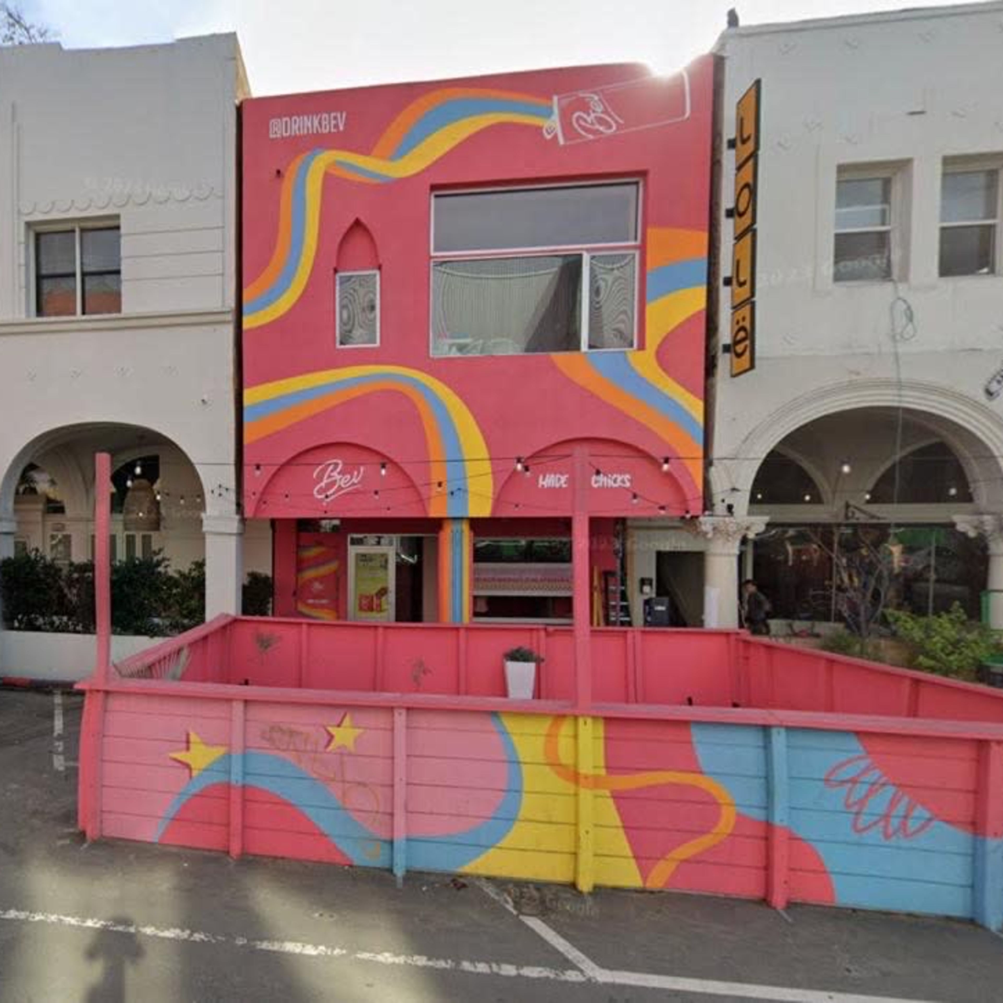 A colorful building with a mural on its exterior, situated between two white structures with arched entries, alongside a parked van and a pink fence.