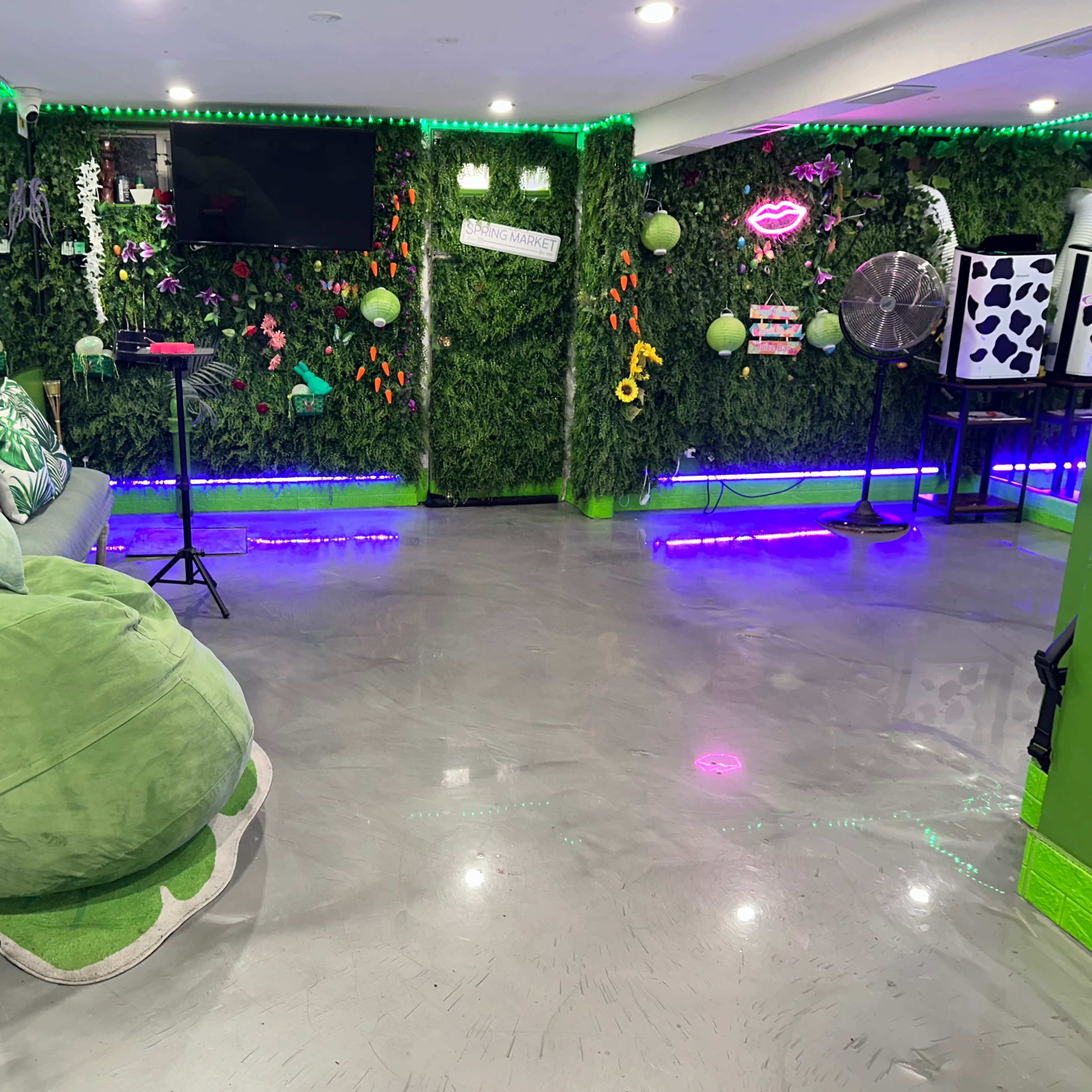 The room features green walls adorned with artificial plants, neon lights, and a variety of decorative elements, including a speaker and a fan.