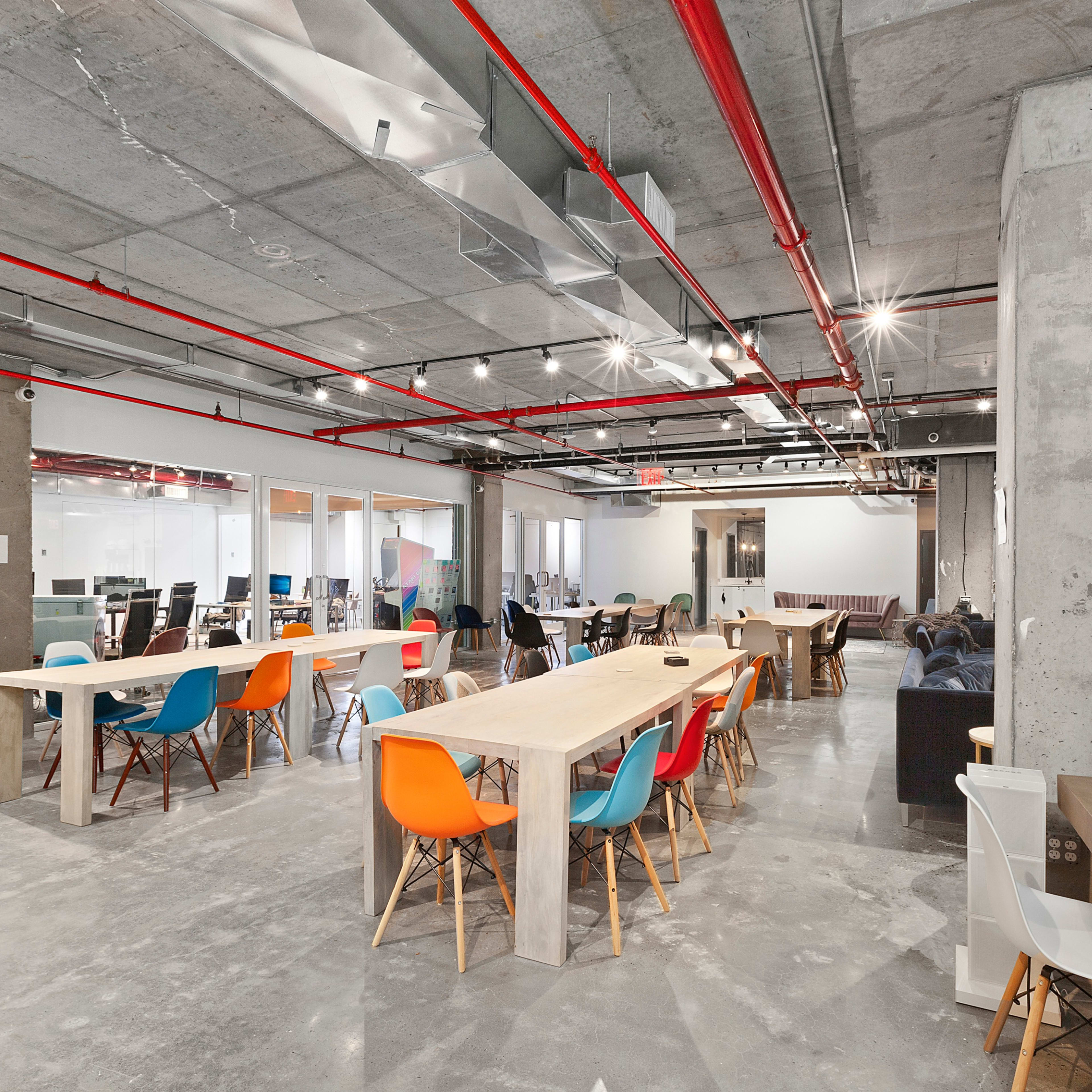 A modern, open office space with long tables, colorful chairs, exposed concrete walls, and visible overhead piping.