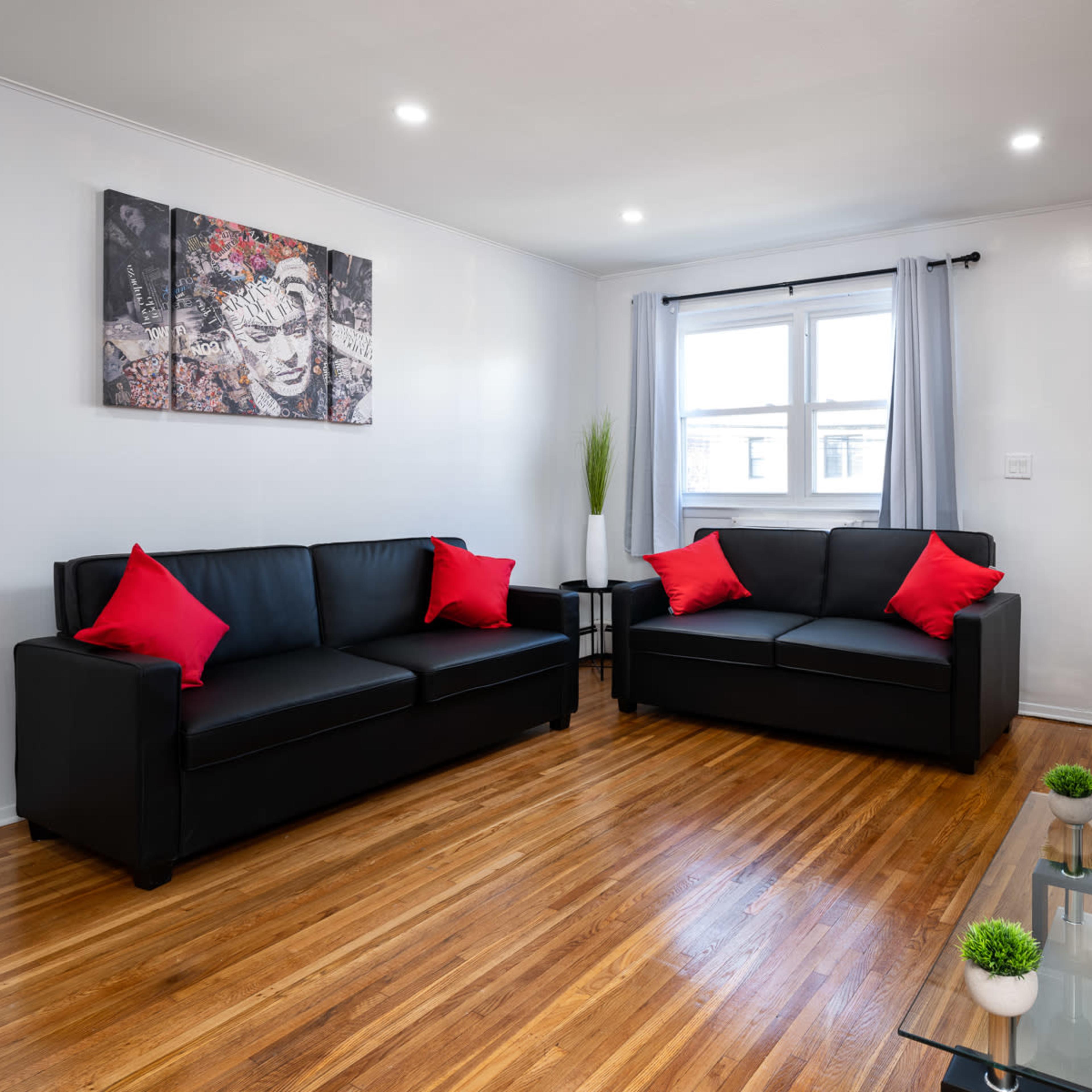 The living room features two black sofas with red pillows, a glass coffee table, and a wall-mounted television, illuminated by natural light from two windows.
