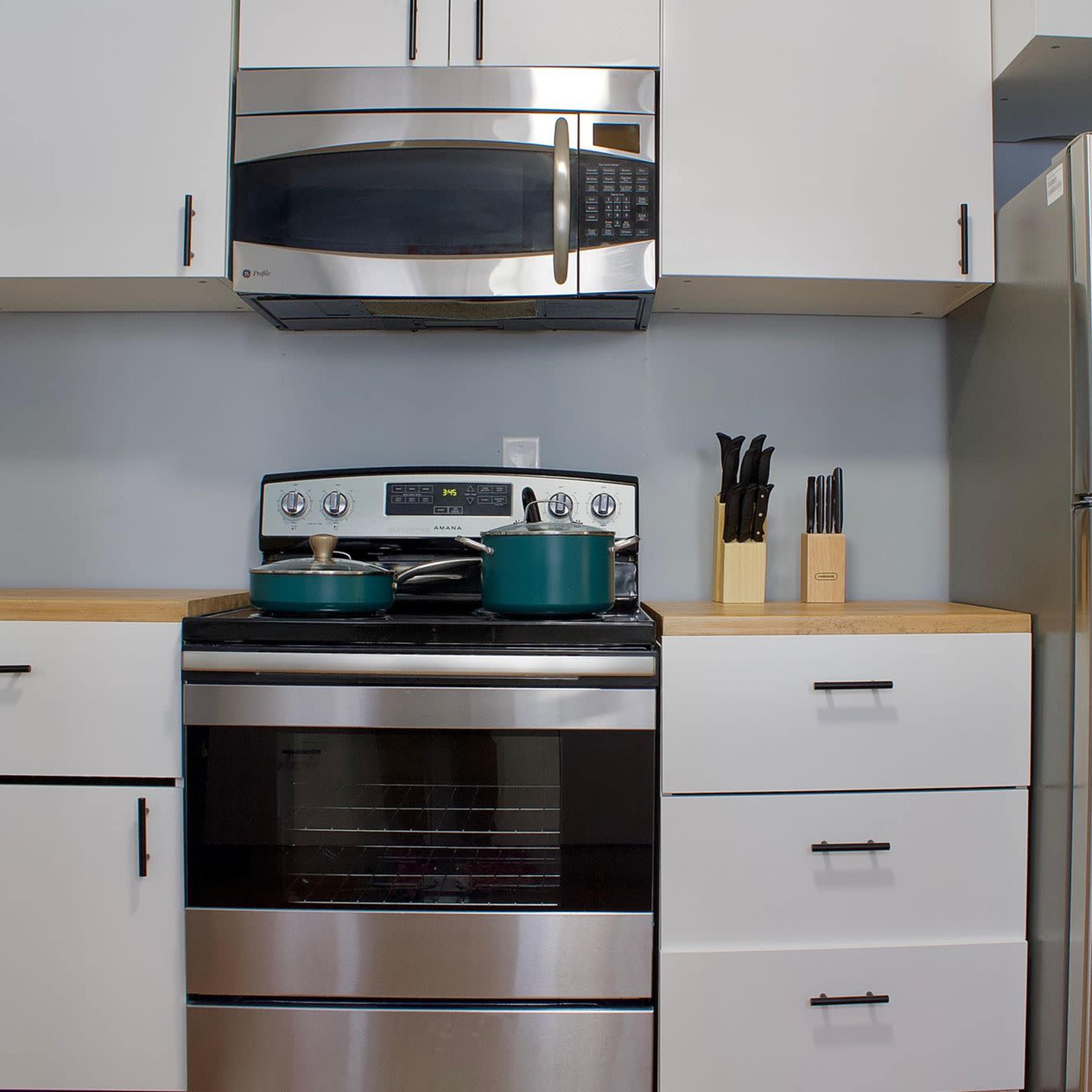 A modern kitchen setup with a stainless steel stove, microwave, and refrigerator, alongside blue cookware on the stovetop and utensils in a holder.