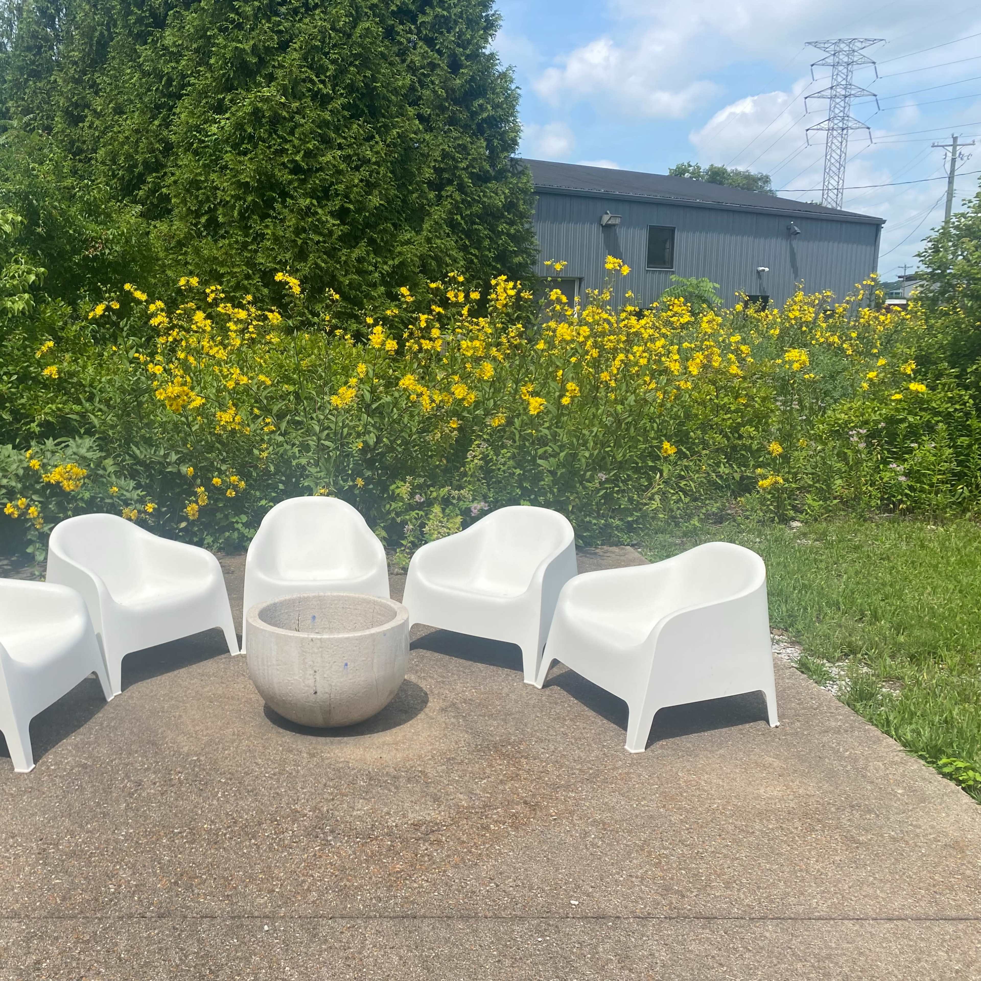 A group of five white chairs is arranged in a circle around a central stone table, set against a backdrop of yellow flowers and greenery.