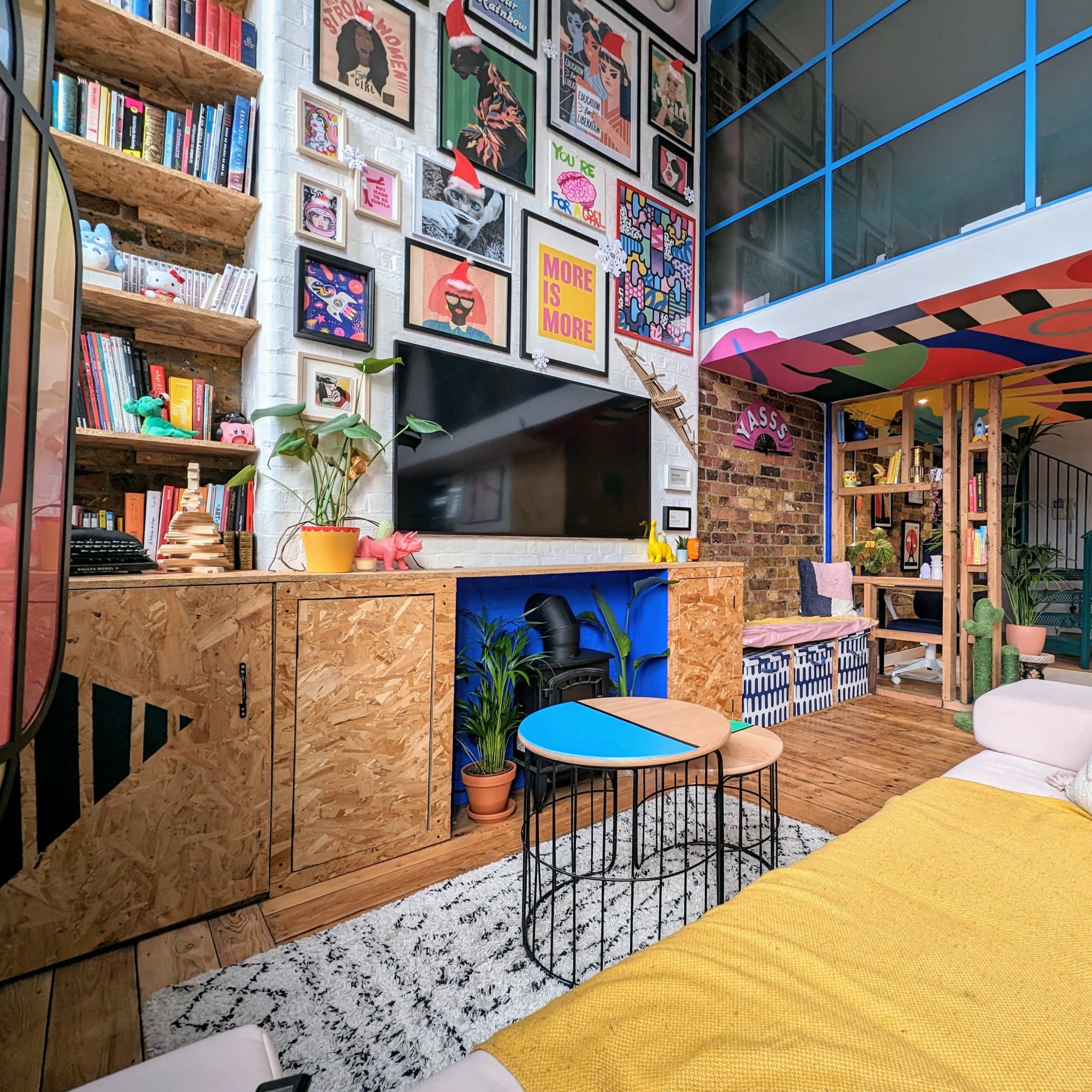 A colorful living room with a modern design, showcasing a wall adorned with framed artwork, a cozy seating area, and shelves filled with books and decorative items.