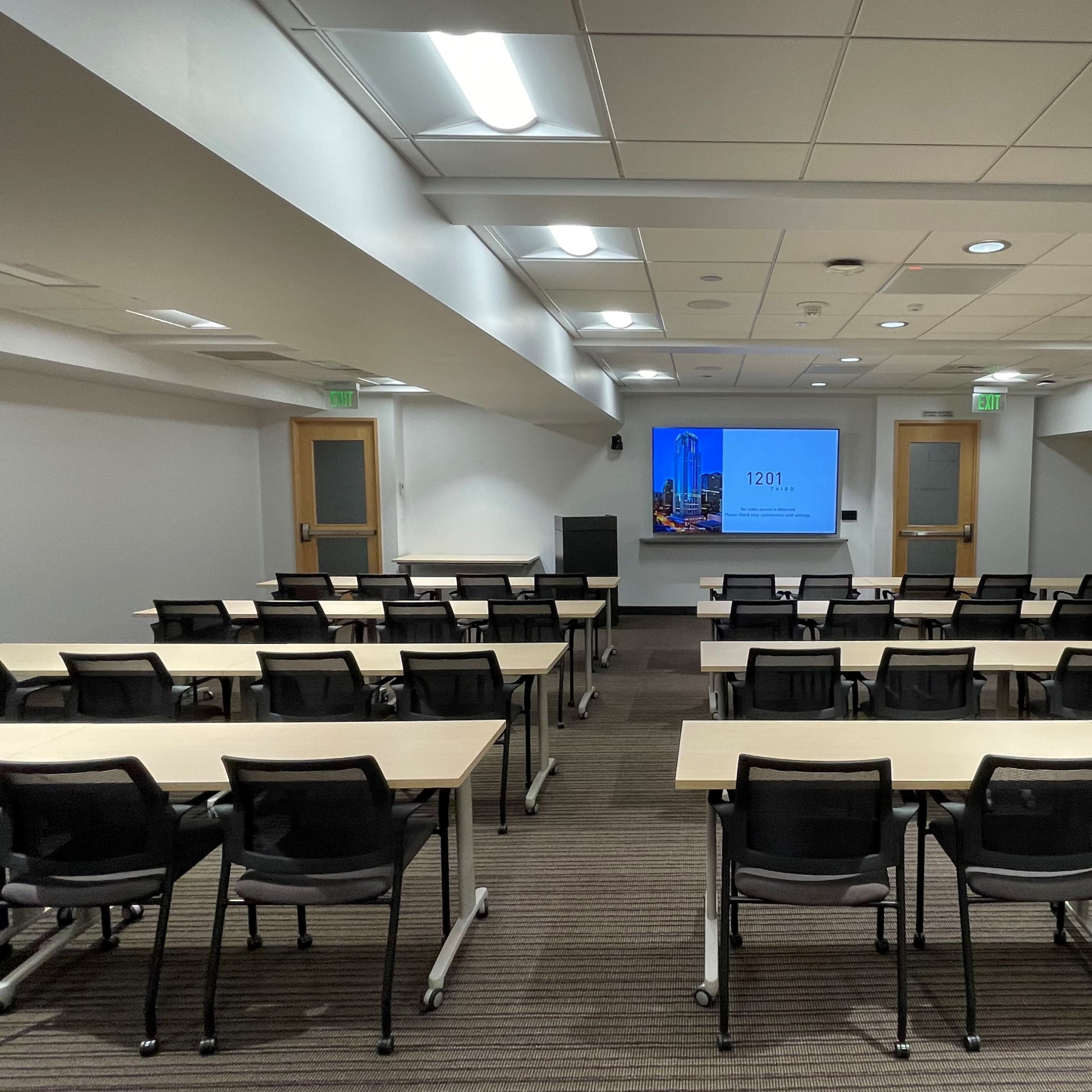 A conference room with rows of tables and chairs facing a screen displaying a presentation, illuminated by overhead lighting.