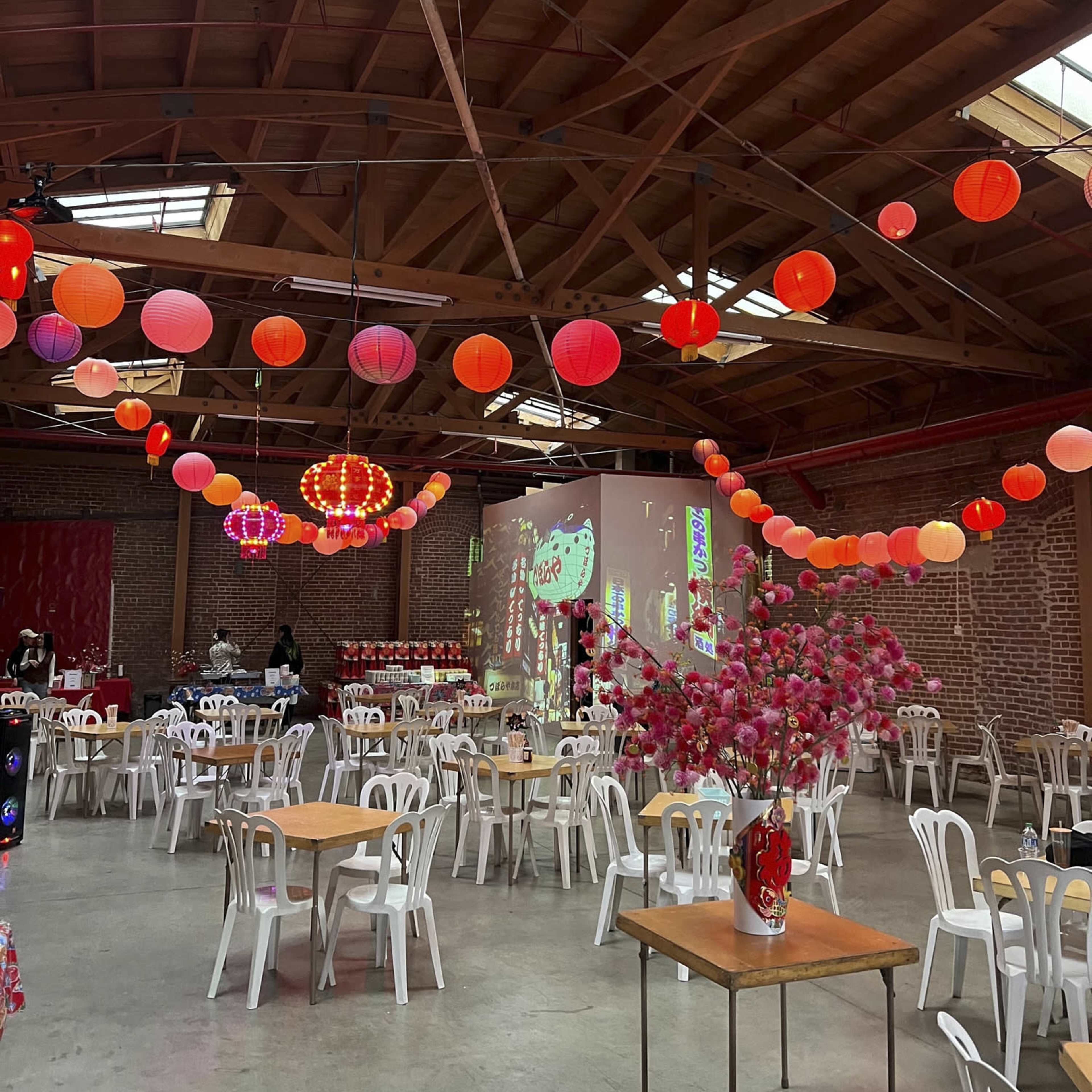A spacious indoor venue with wooden beams, tables and chairs arranged, and colorful paper lanterns hanging from the ceiling.