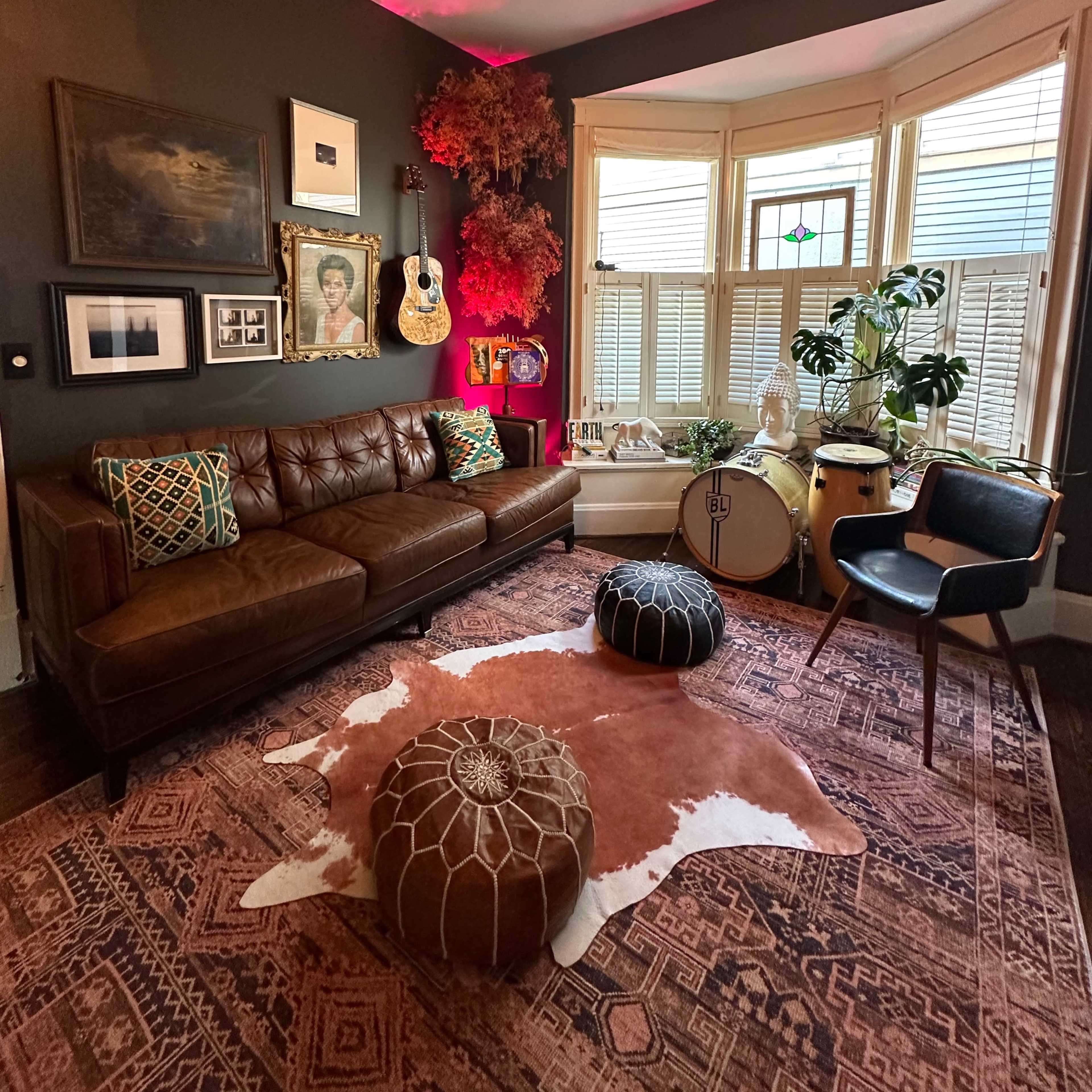 A cozy living room features a brown leather sofa, decorative poufs, a patterned rug, and a bay window filled with plants and art.