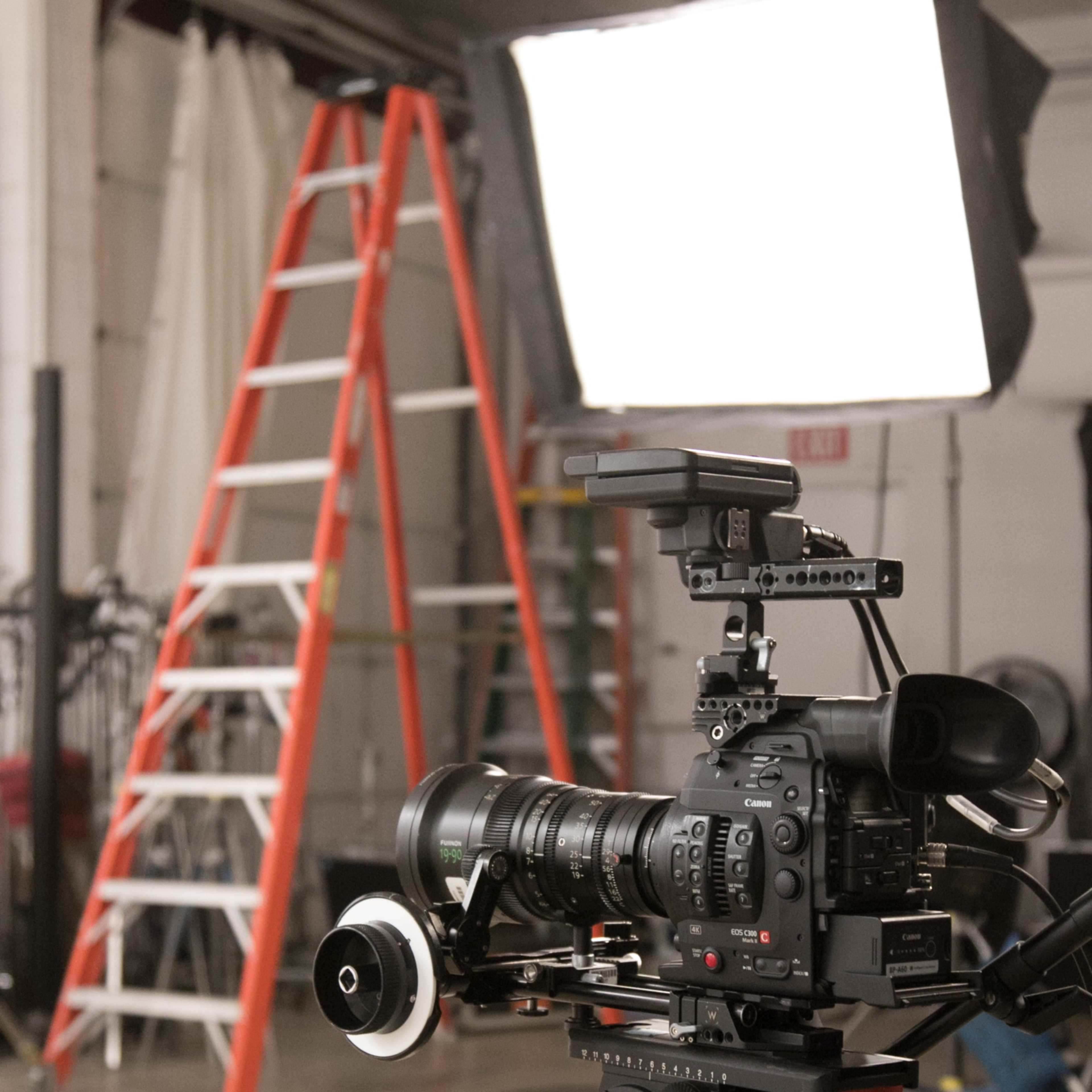 A professional video camera is set on a tripod in a studio with an illuminated softbox and a ladder in the background.