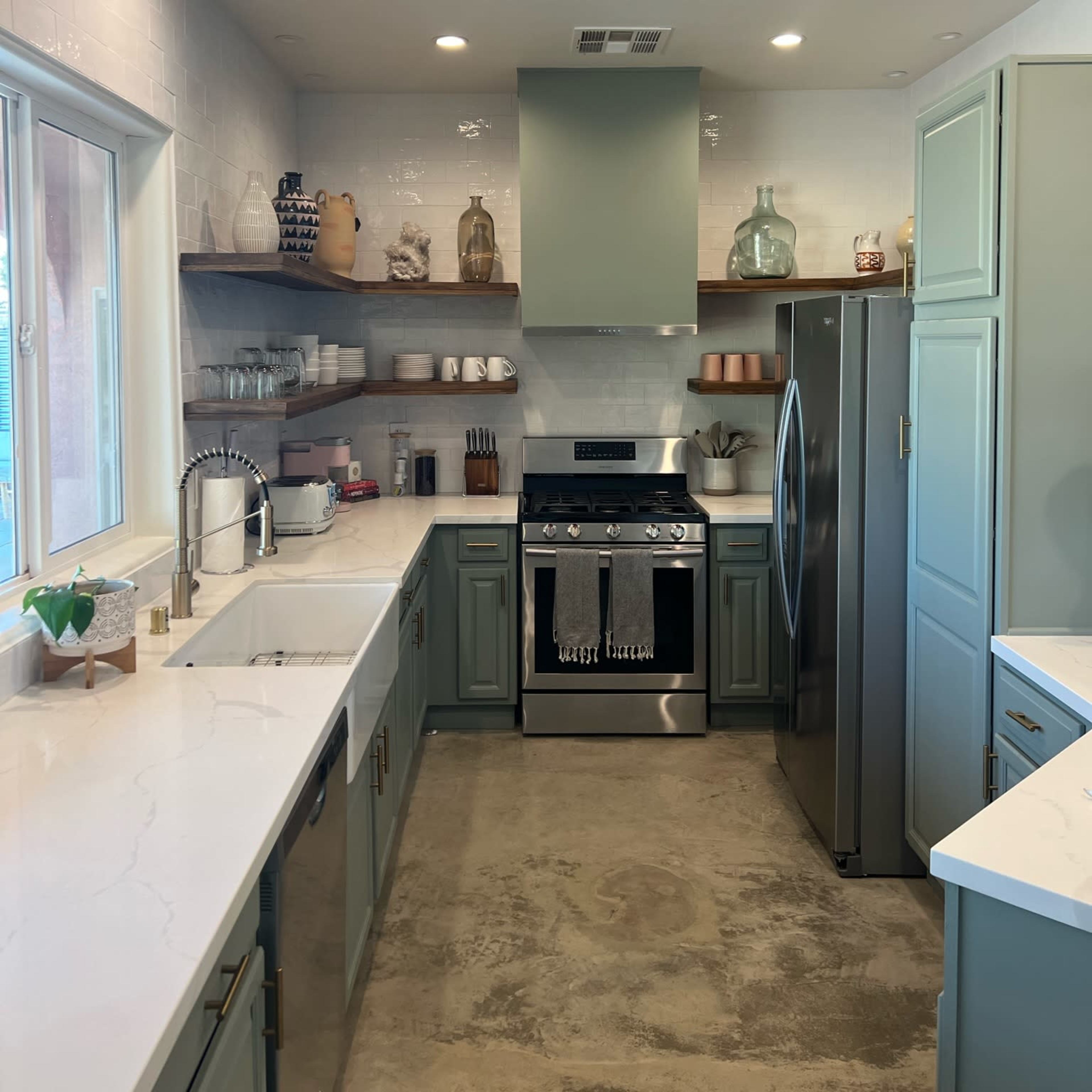 A modern kitchen with green cabinets, a silver refrigerator, a stove, and shelves displaying various kitchen items.