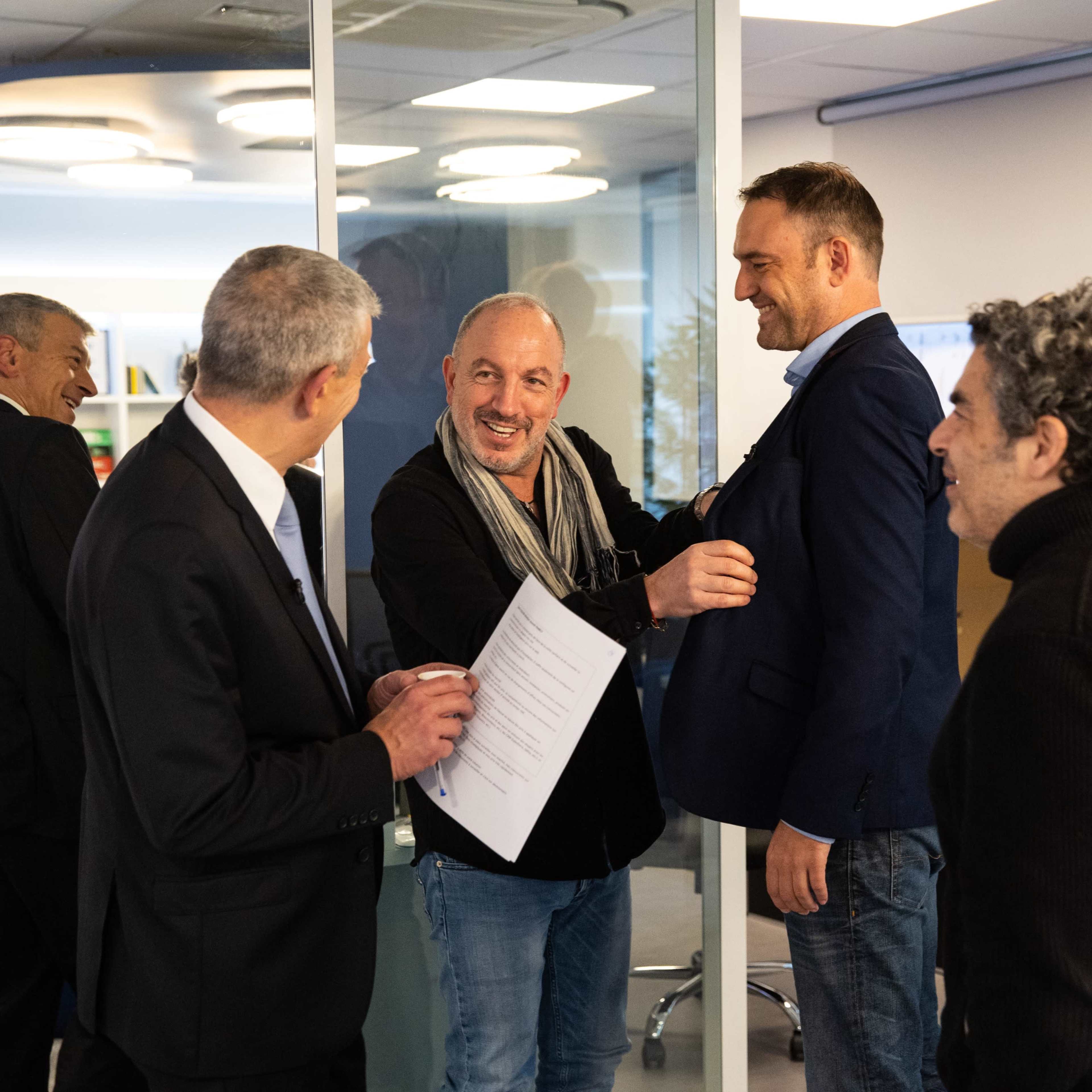 A group of men is interacting in an office setting, with one man adjusting the jacket of another while holding a document.