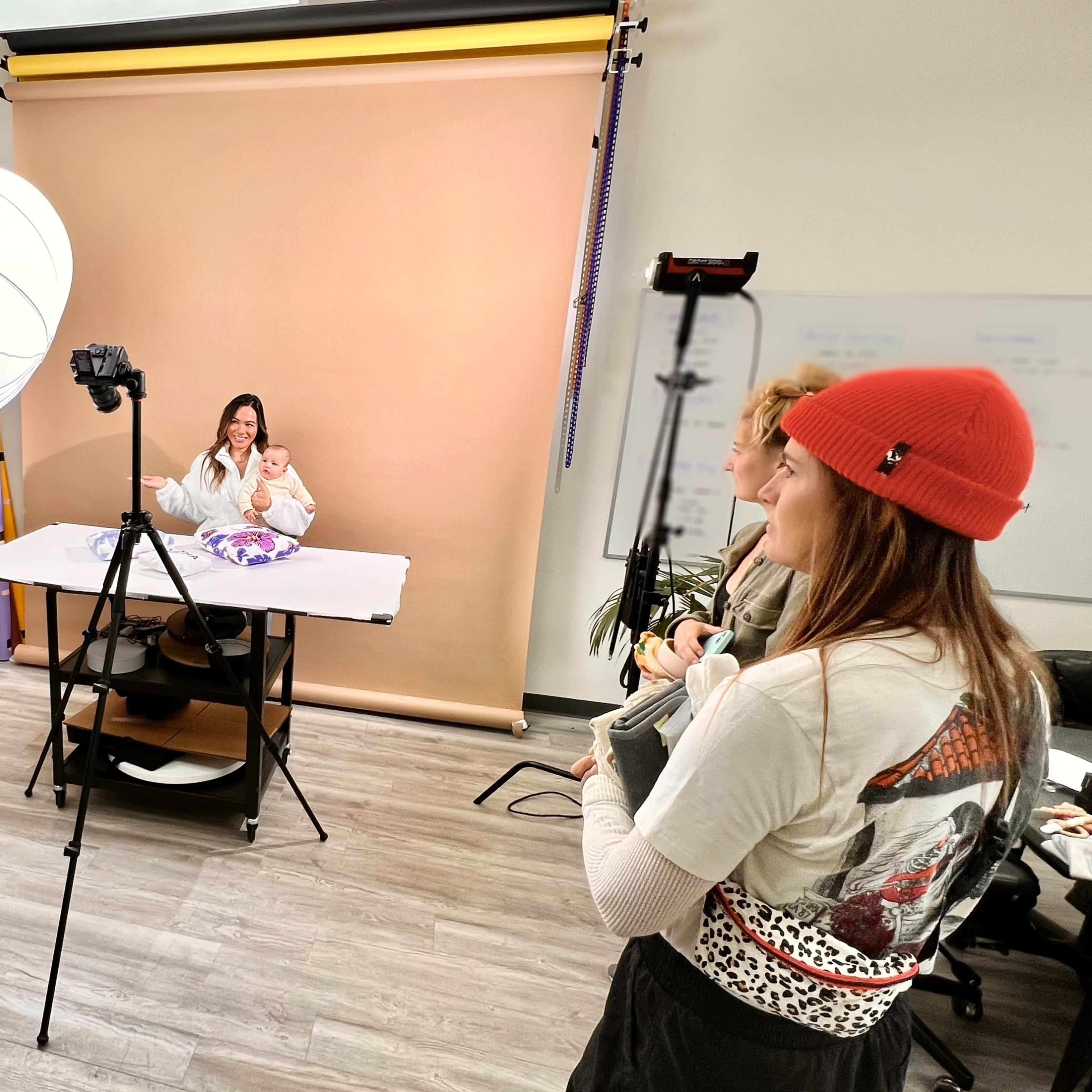 A woman sits at a table in front of a camera, while two people stand nearby, one holding a notebook and another adjusting a light.