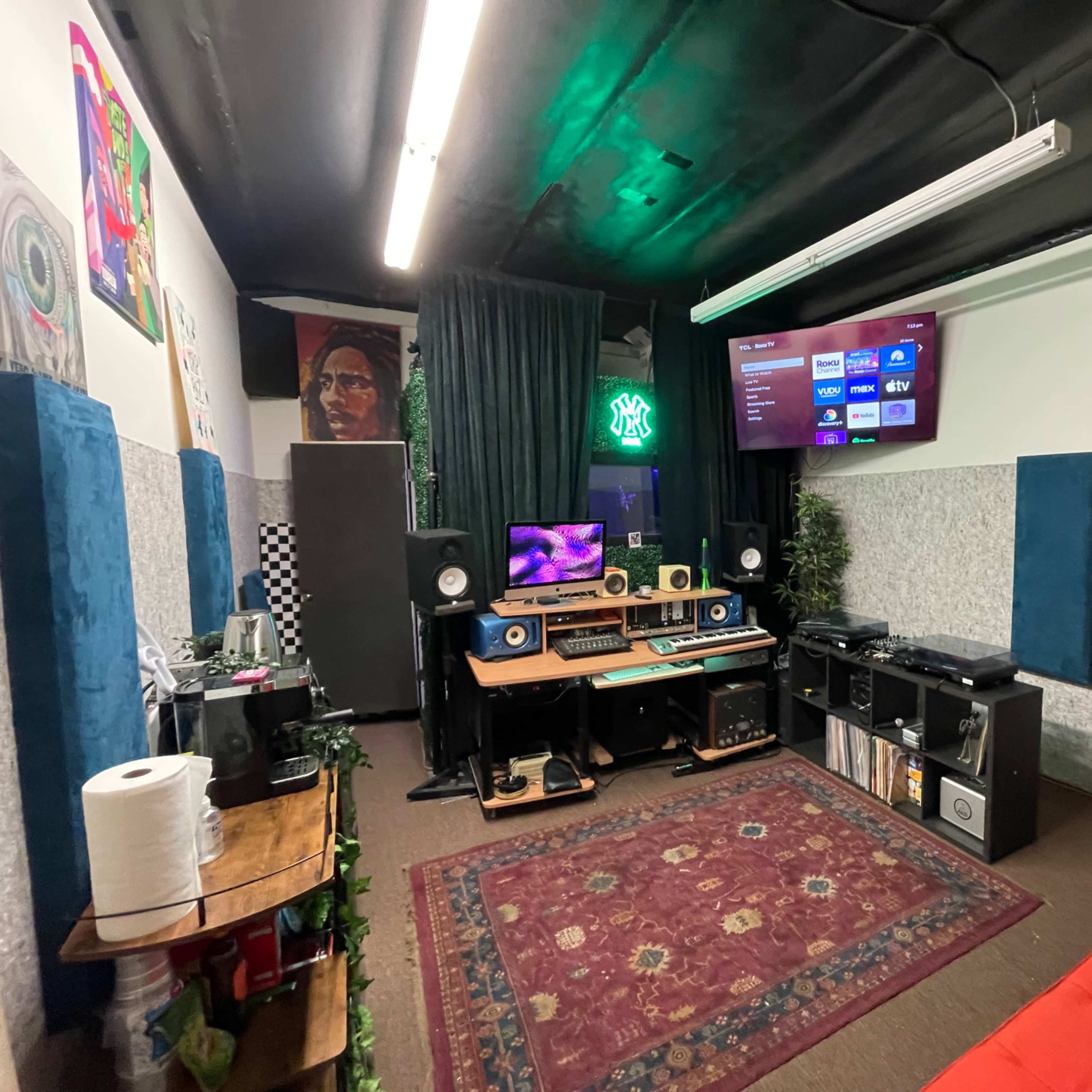 A recording studio with a large mixing desk, speakers, a television, and various musical equipment surrounded by colorful artwork and decorative elements.