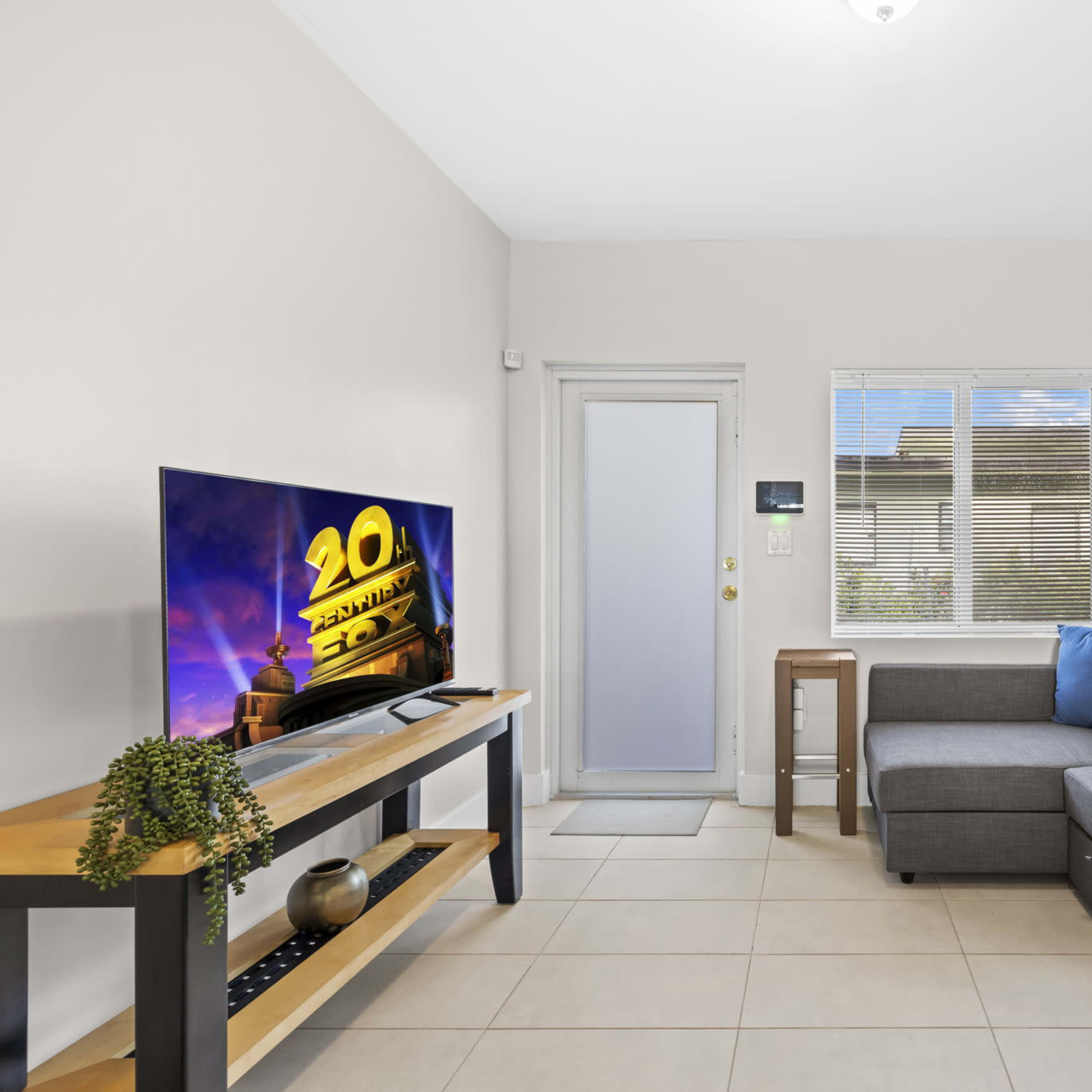 A living room with a gray sectional sofa, a television on a wooden stand, and a door leading outside, accompanied by framed artwork on the wall.