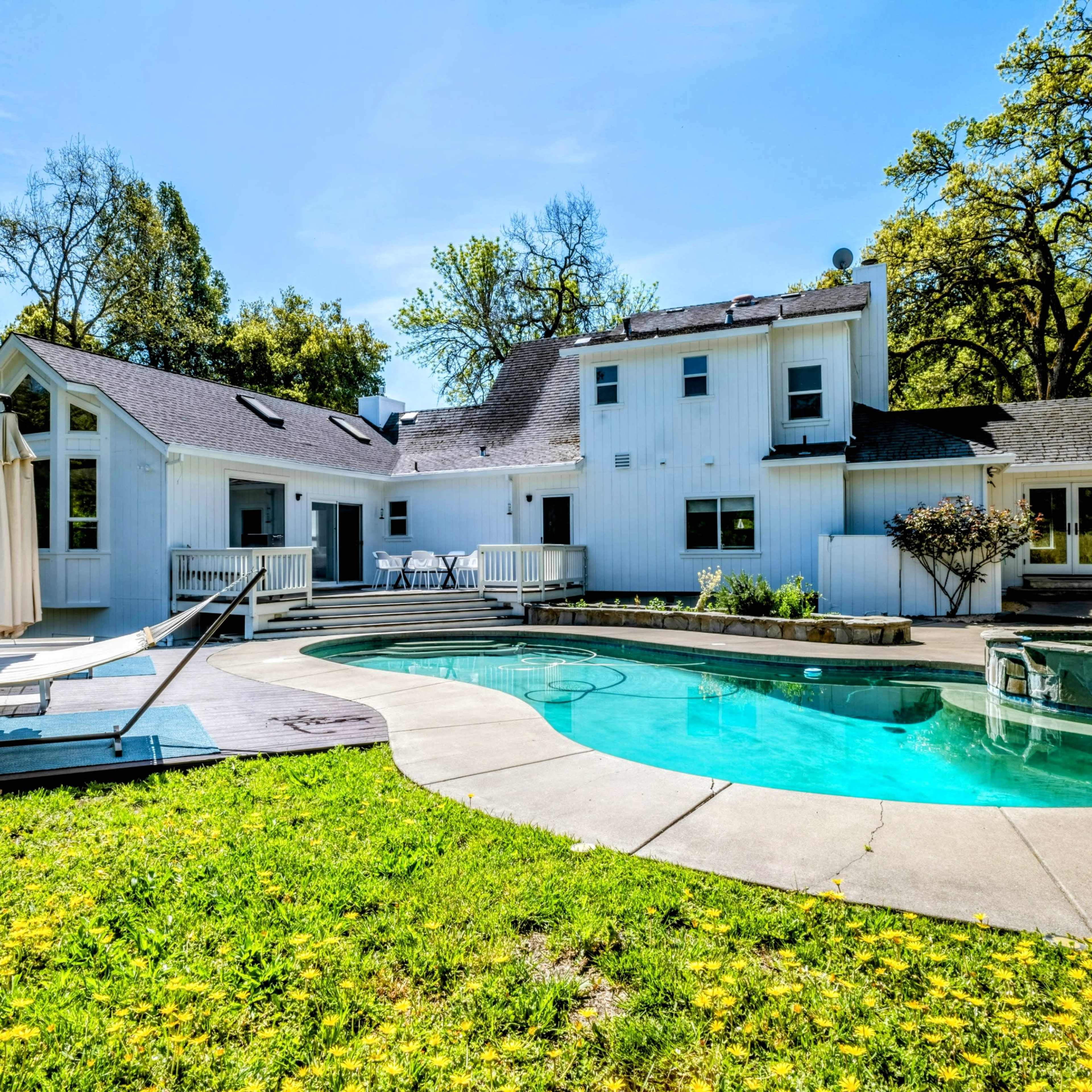 A white house with a swimming pool and lounge chairs in a grassy backyard surrounded by trees.