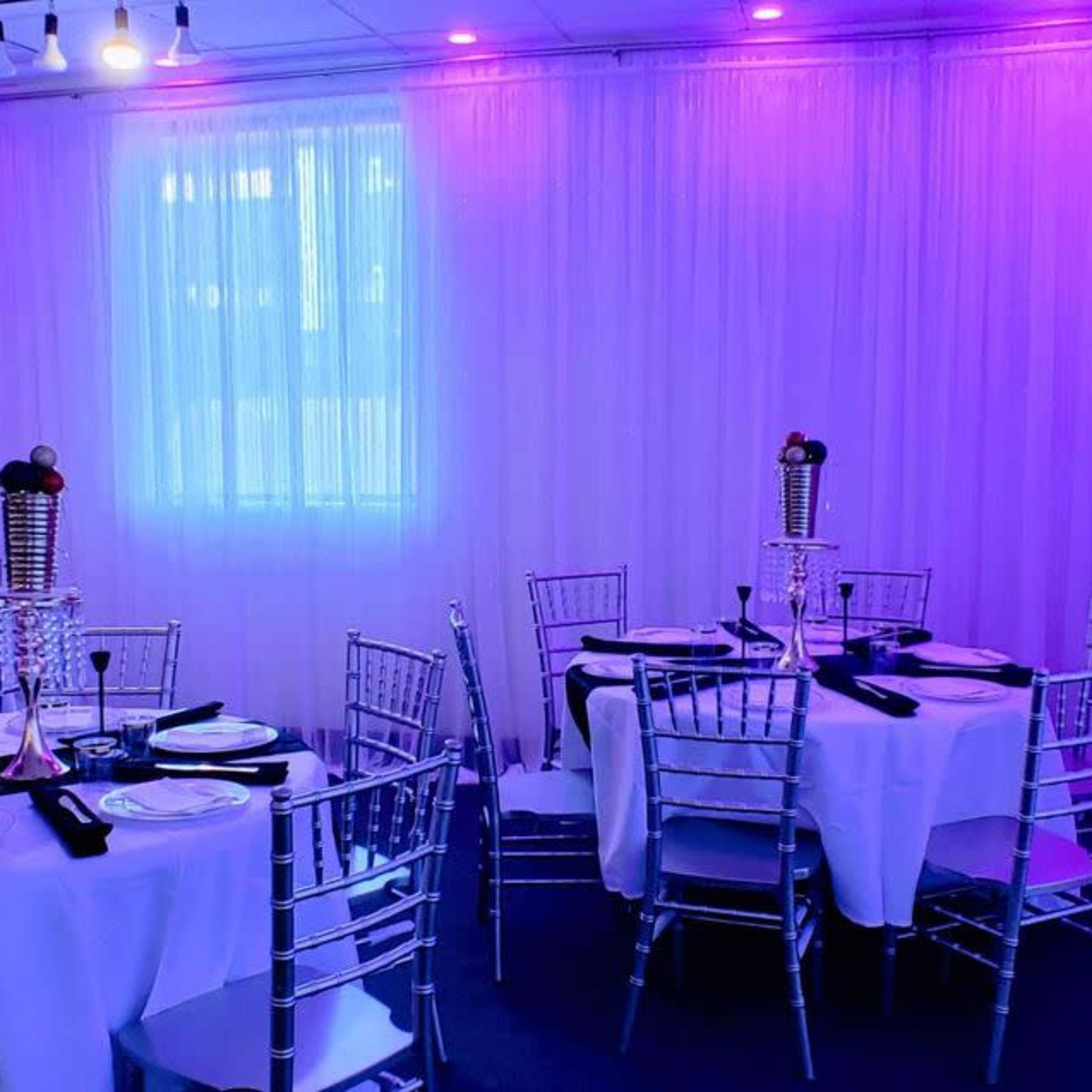 A dining area features round tables set with white tablecloths, silver chairs, and decorative centerpieces under purple lighting.