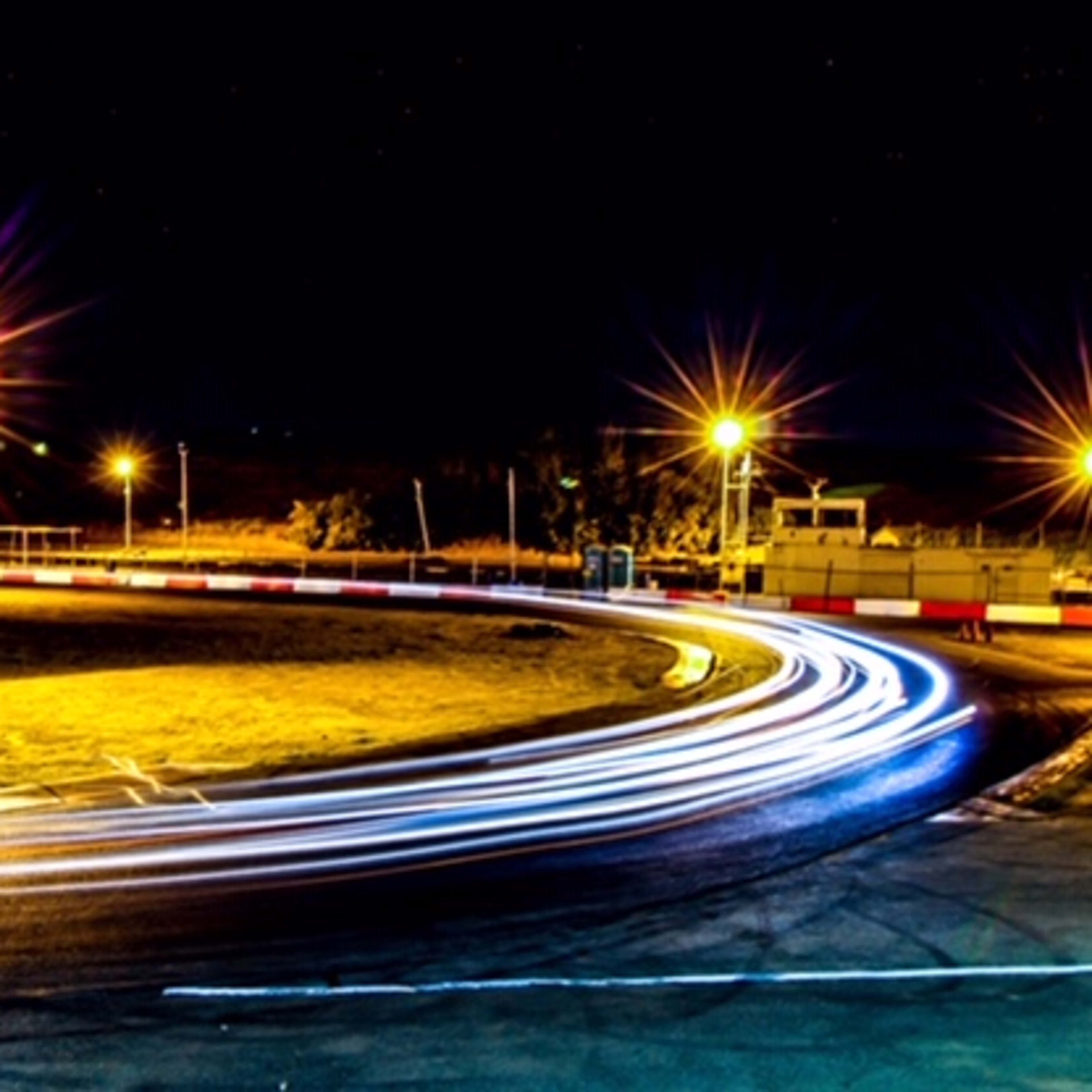 A race track illuminated by bright lights, with streaks of car lights following a curved path at night.