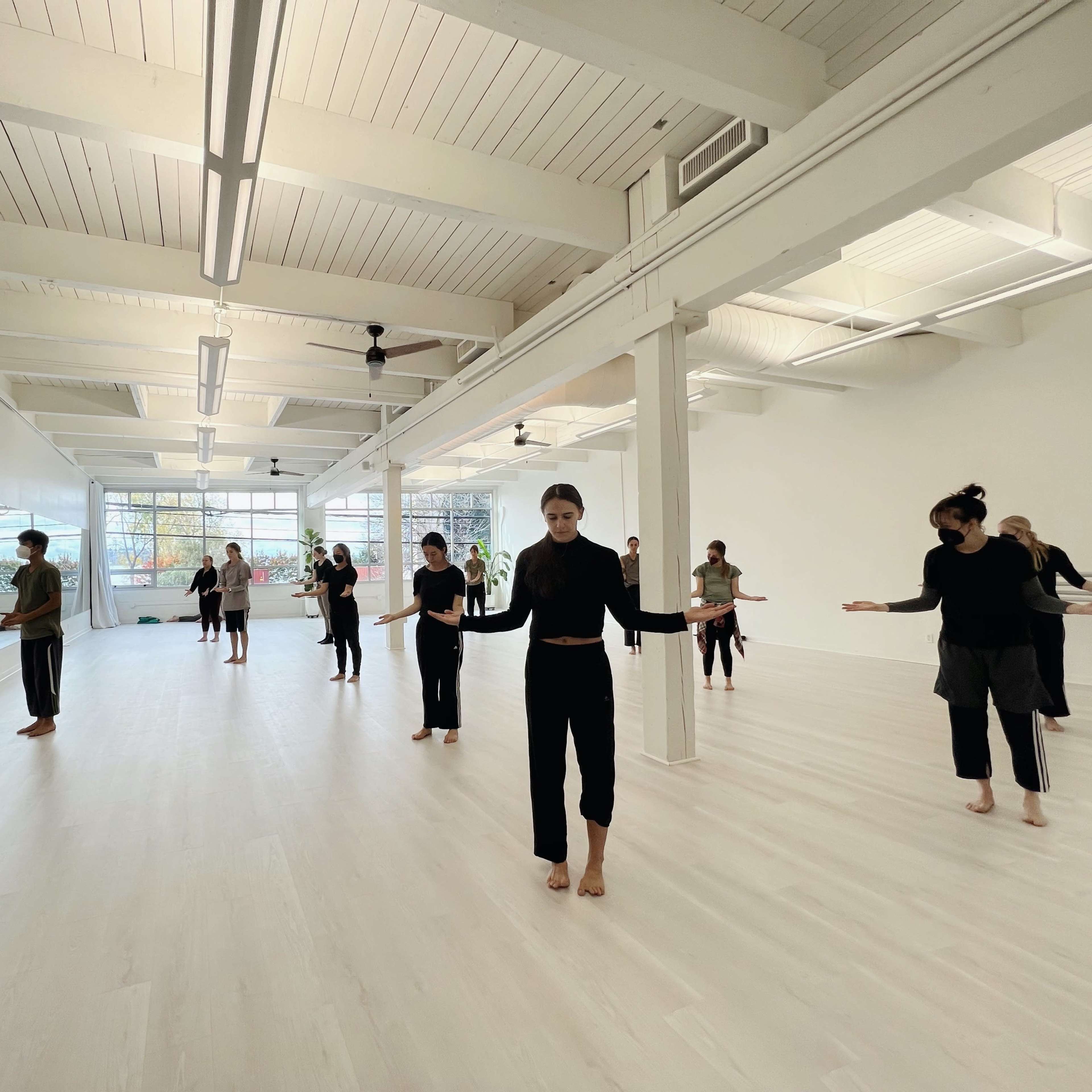A group of people is practicing movement in a spacious, well-lit studio with large mirrors.