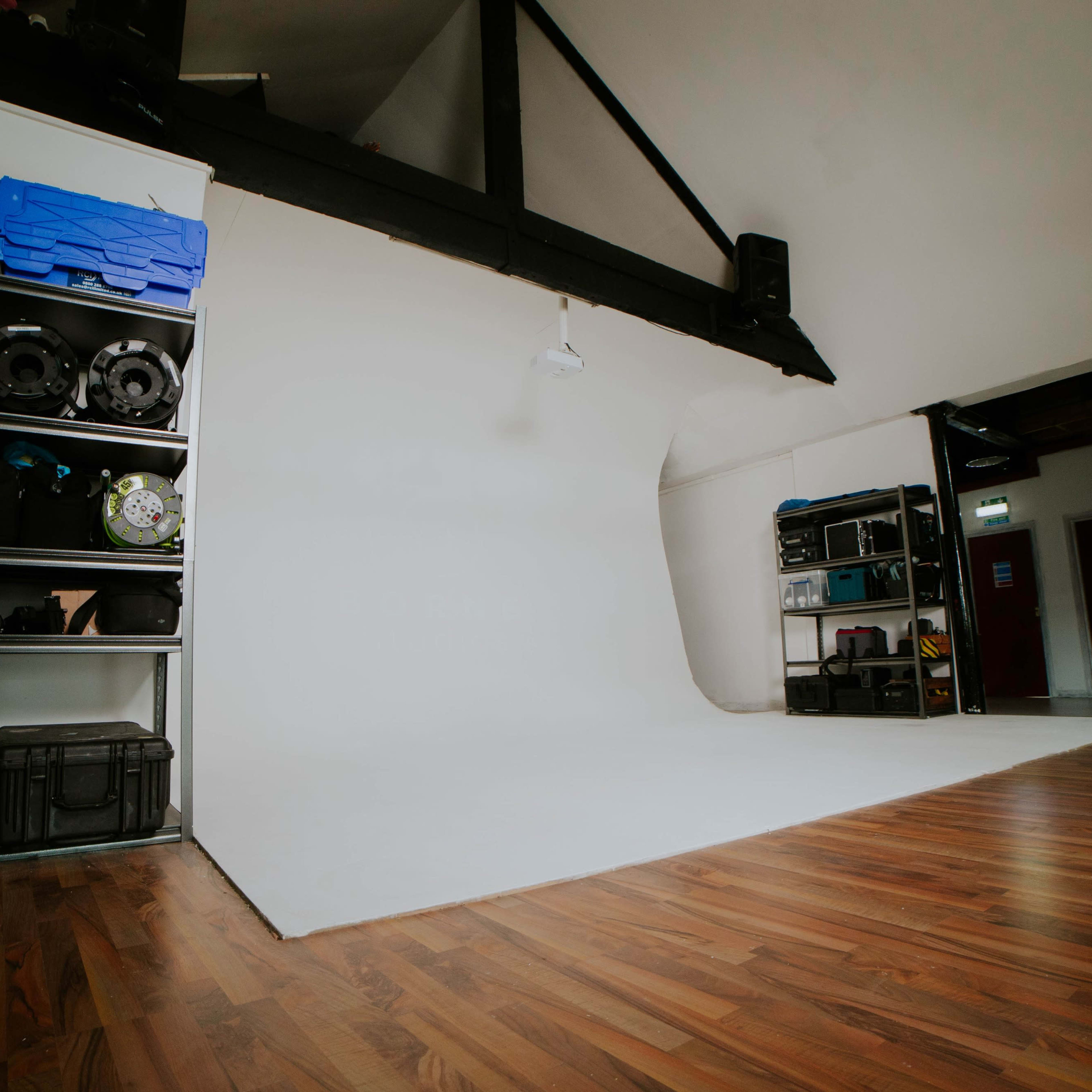 A photography studio with a white backdrop, wooden flooring, and shelves containing various equipment on one side.