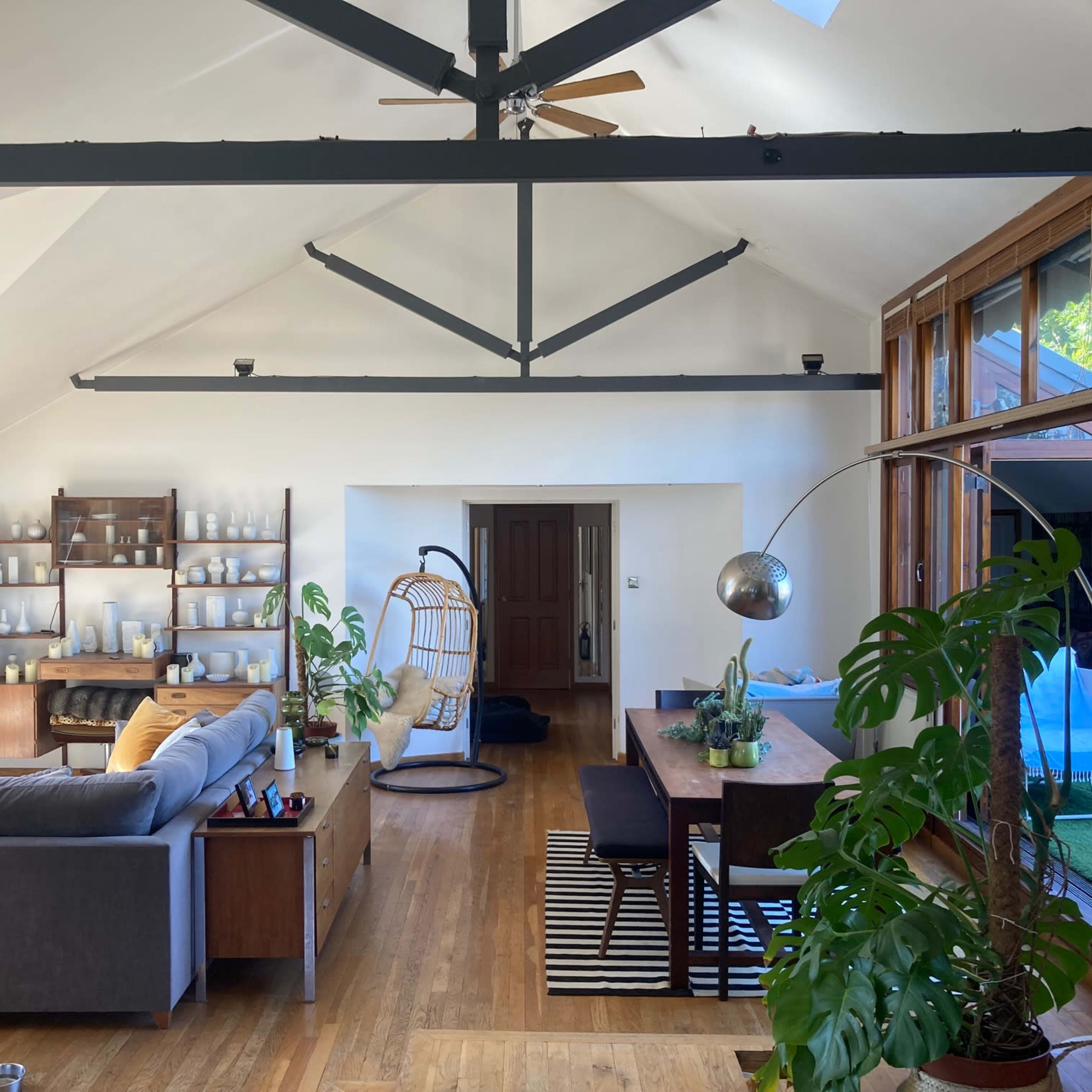 A spacious, modern living area featuring a gray sofa, dining table, and a hanging chair, with large windows allowing natural light to fill the space.