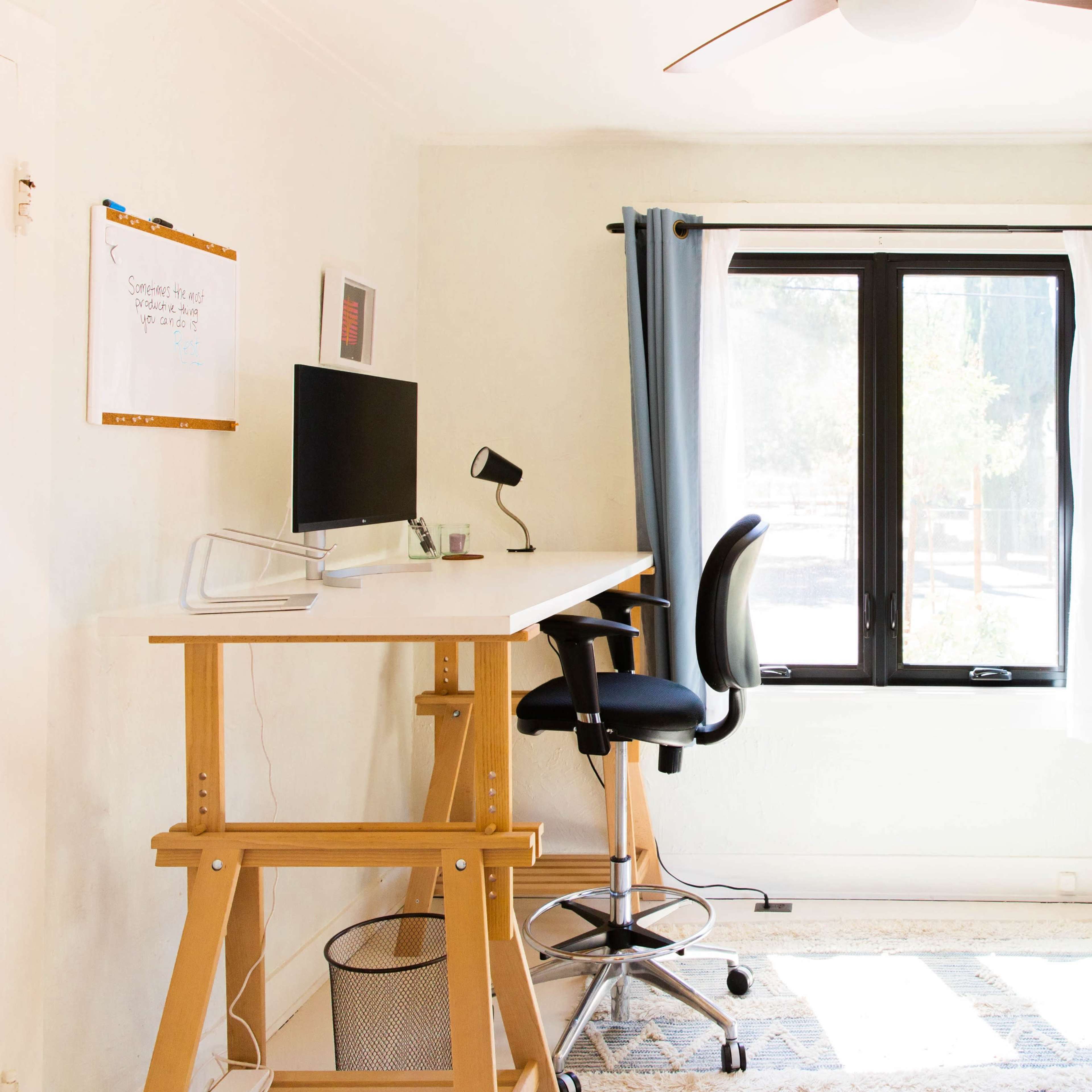 A small home office features a desk with a computer, a rolling chair, and a window with blue curtains.