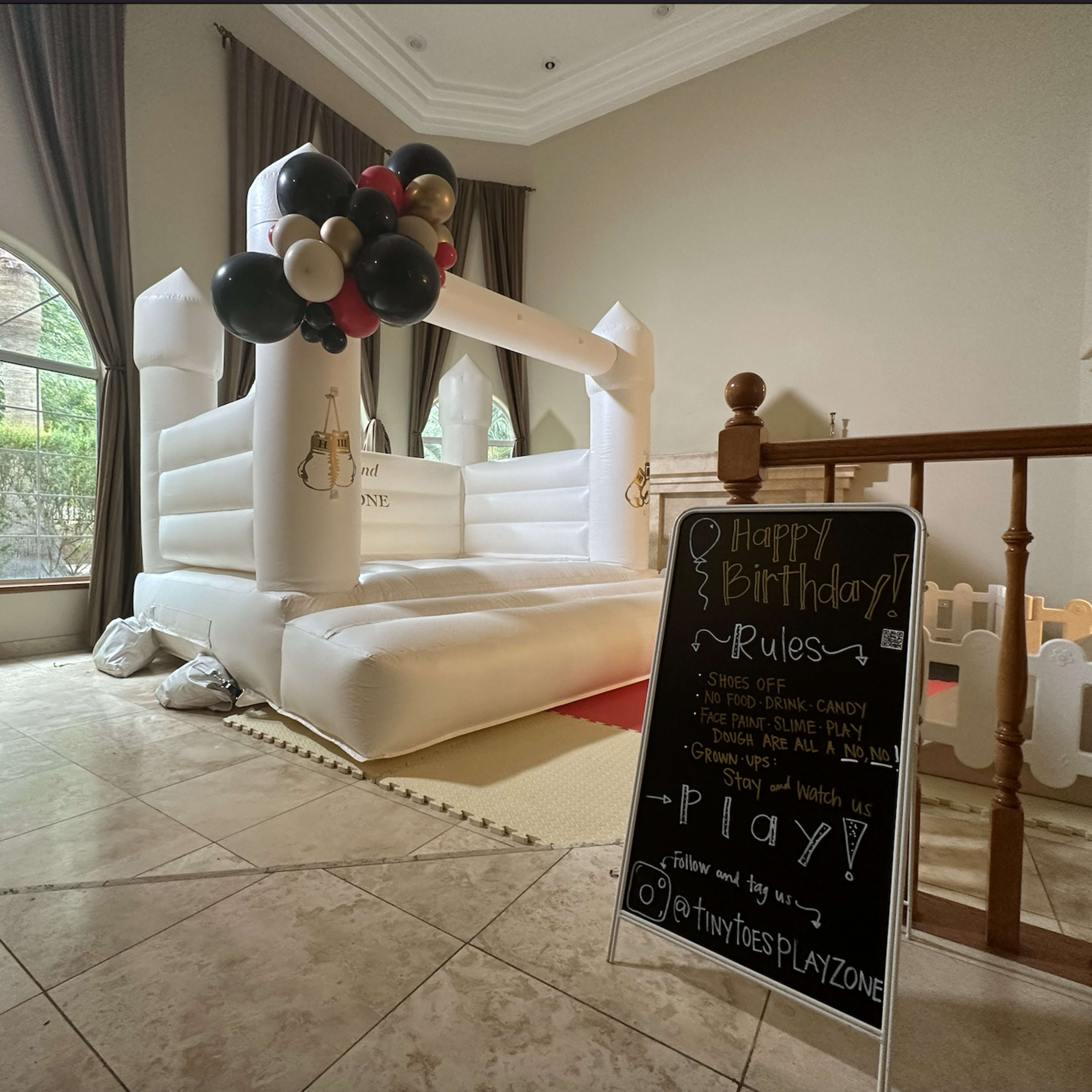 A white inflatable bounce house decorated with black and gold balloons, accompanied by a chalkboard displaying birthday party rules nearby.