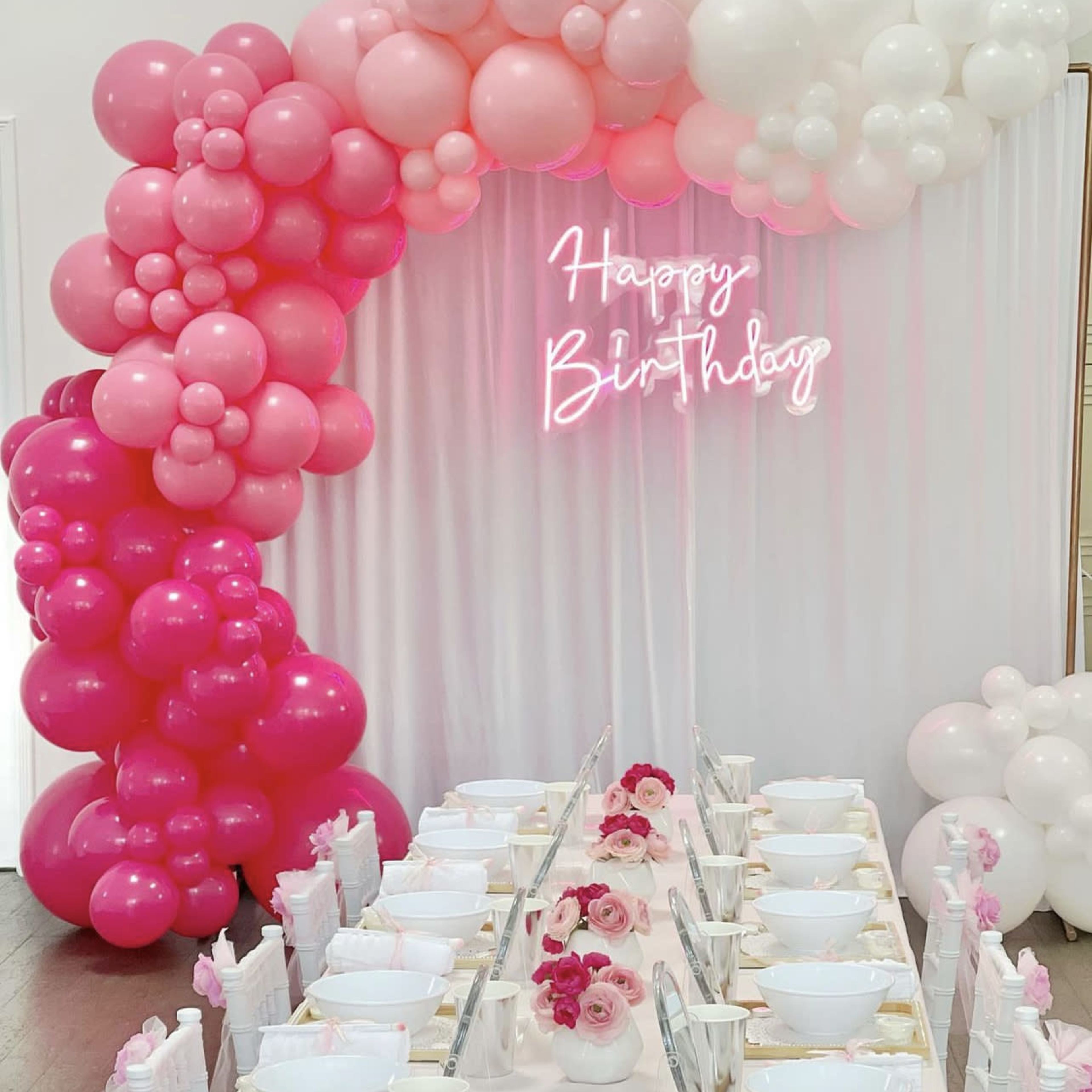 A table is set for a birthday celebration, with a decorative balloon arch in shades of pink and white and a neon "Happy Birthday" sign.