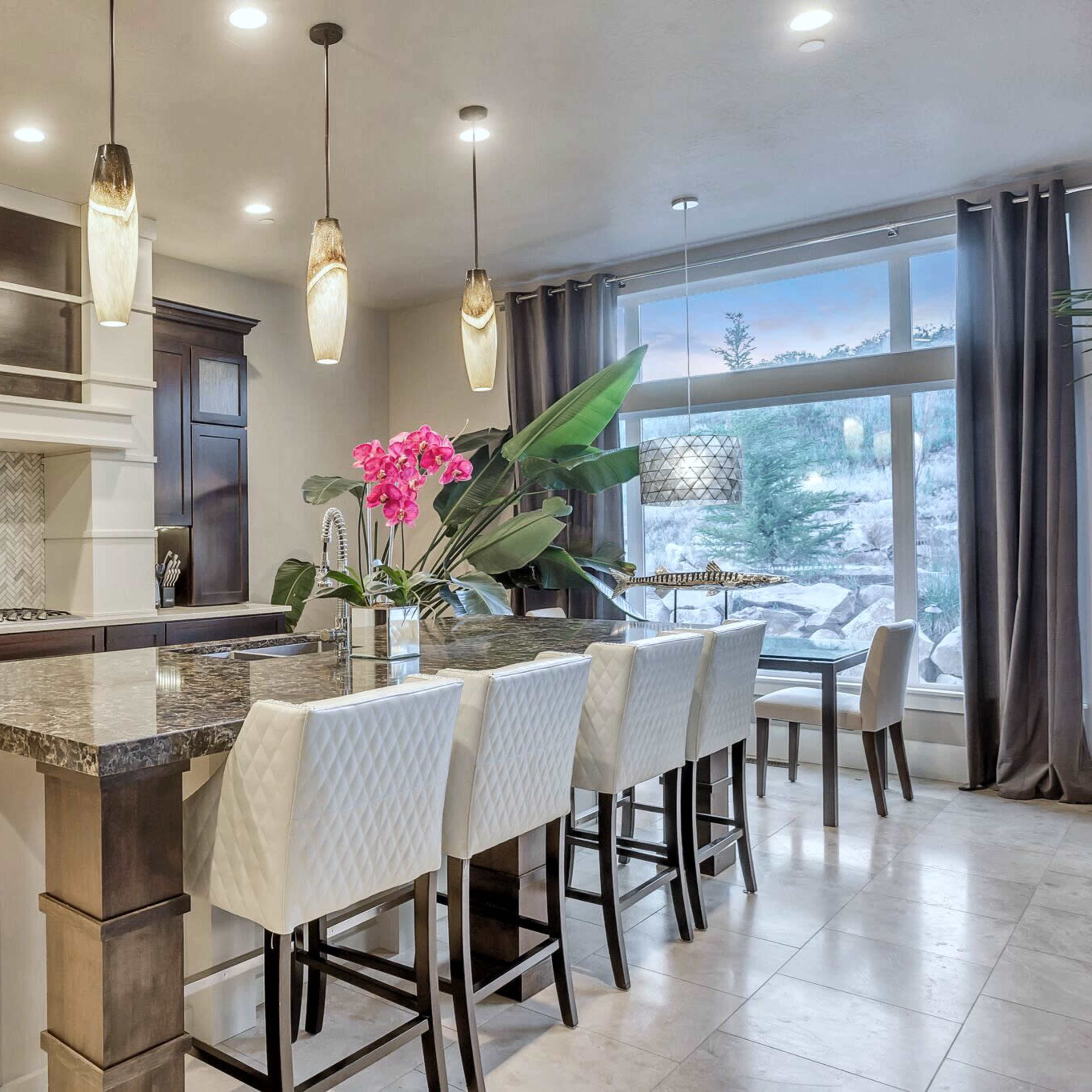 A modern kitchen features dark wood cabinetry, a large island with a granite countertop, and bar seating with white chairs, accented by pink orchids and large windows.