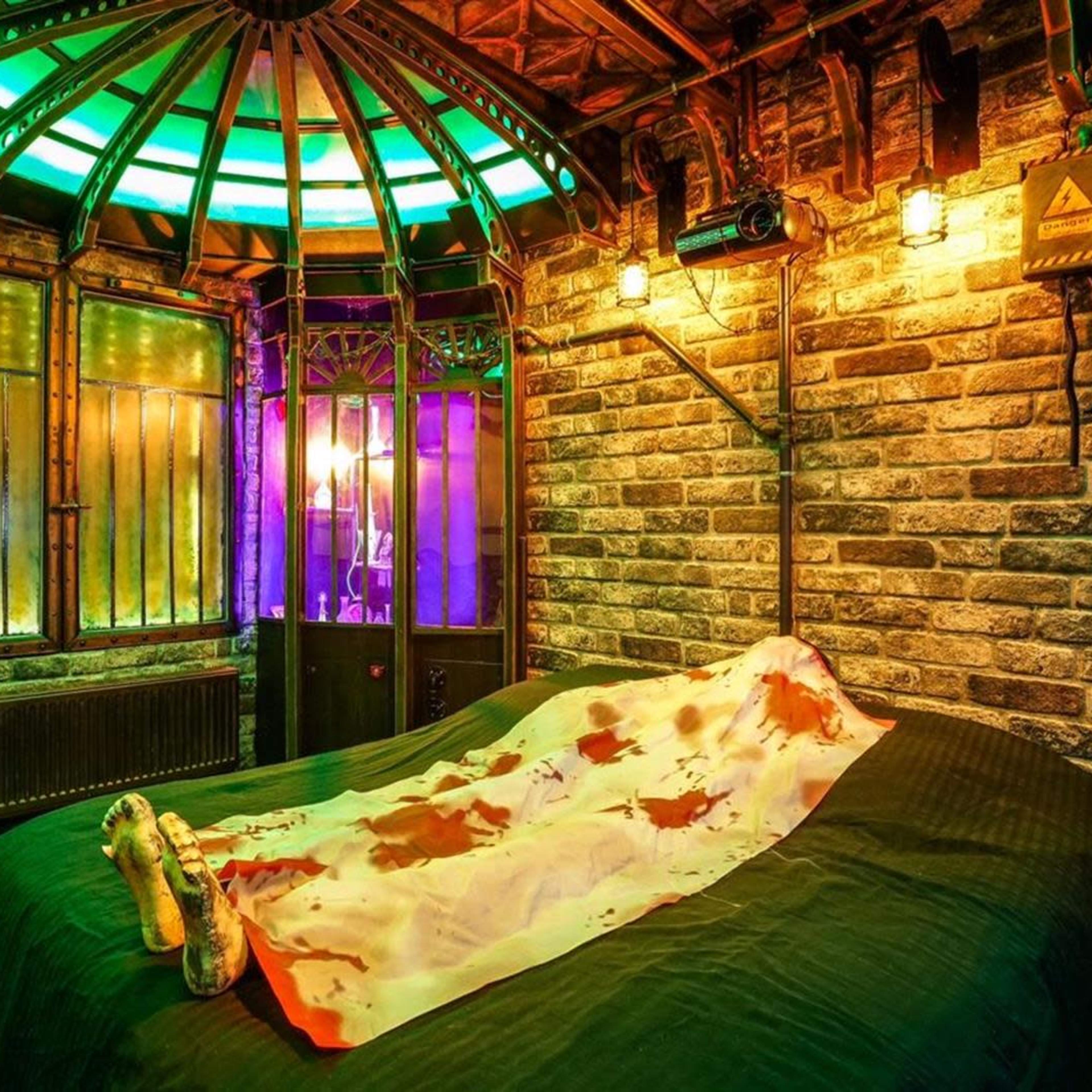 A dimly lit room features a bed with a stained sheet, surrounded by brick walls and a colorful, illuminated ceiling.