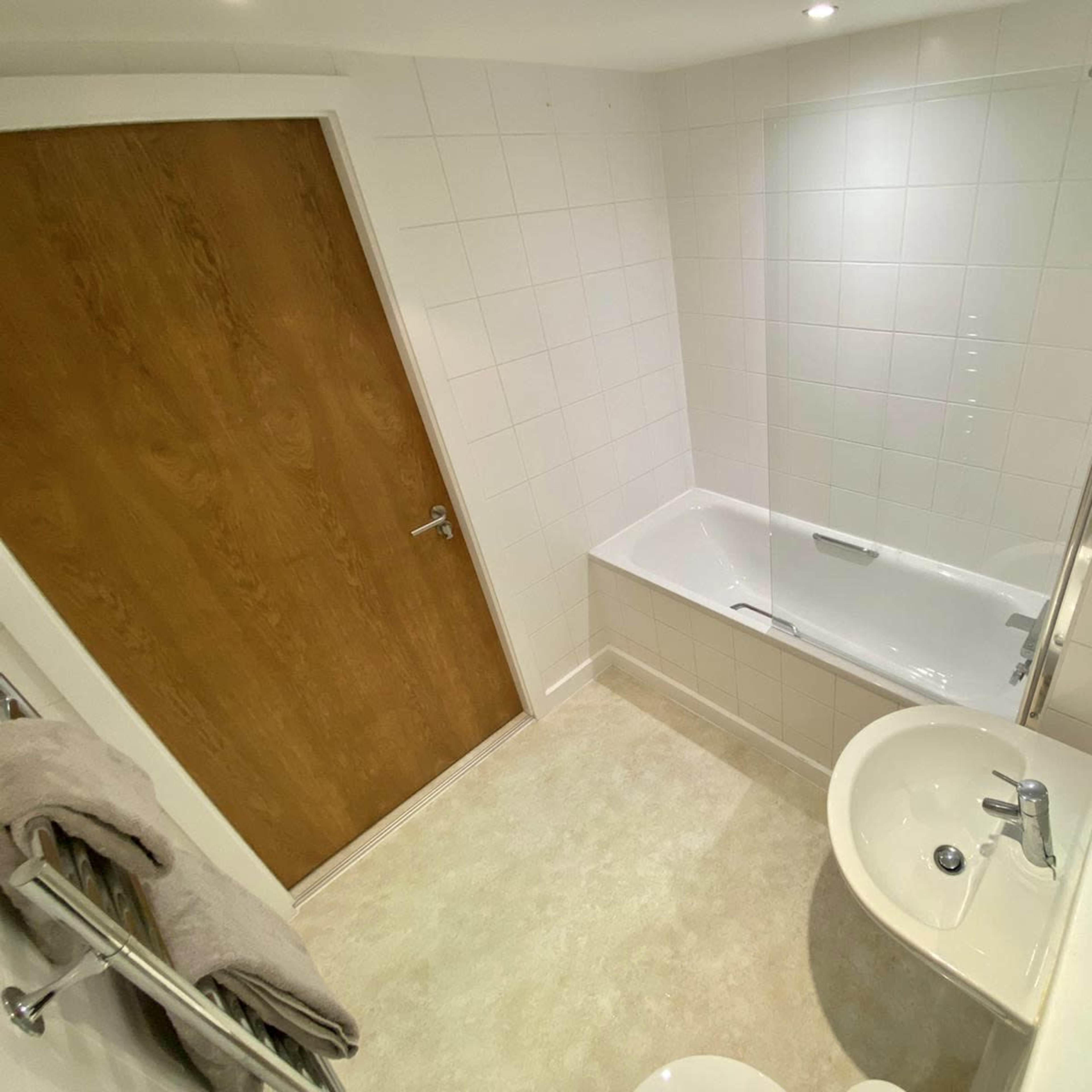 A small bathroom featuring a bathtub with a glass screen, a sink, a mirror, and a wooden door.