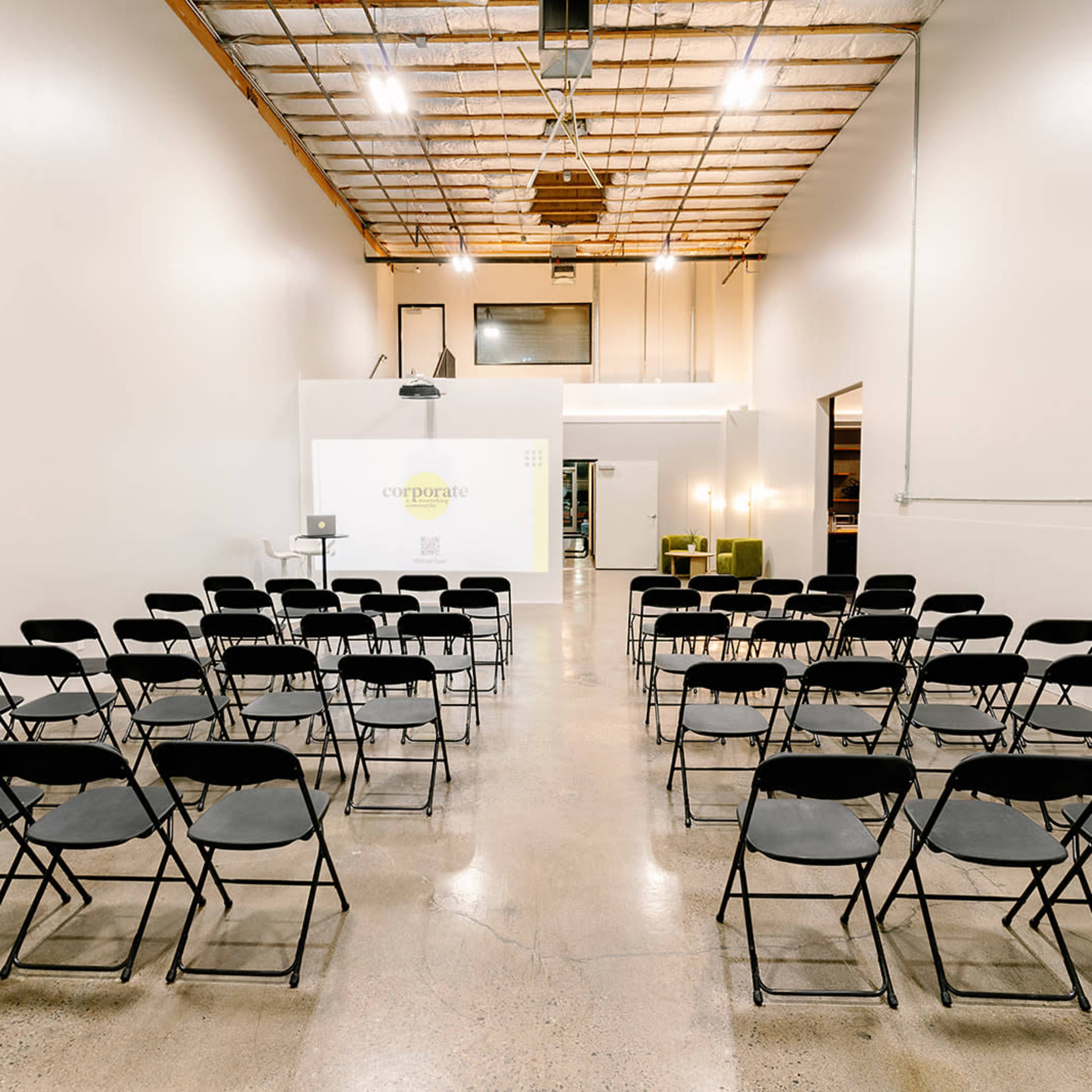 A spacious room with rows of black folding chairs facing a projector screen and a white wall.