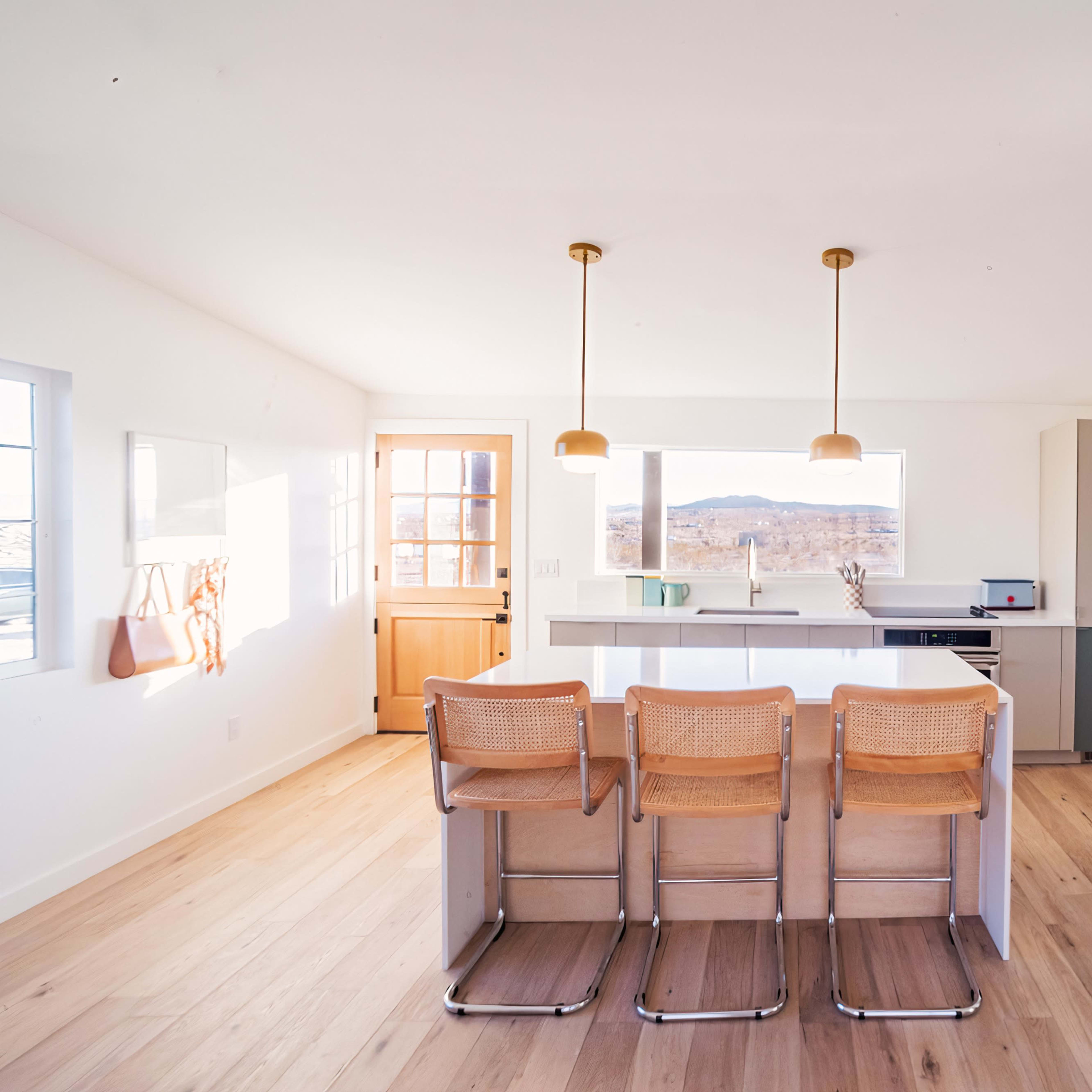 A modern kitchen with a light color scheme, featuring a central island with three bar stools, large windows, and a door leading outside.