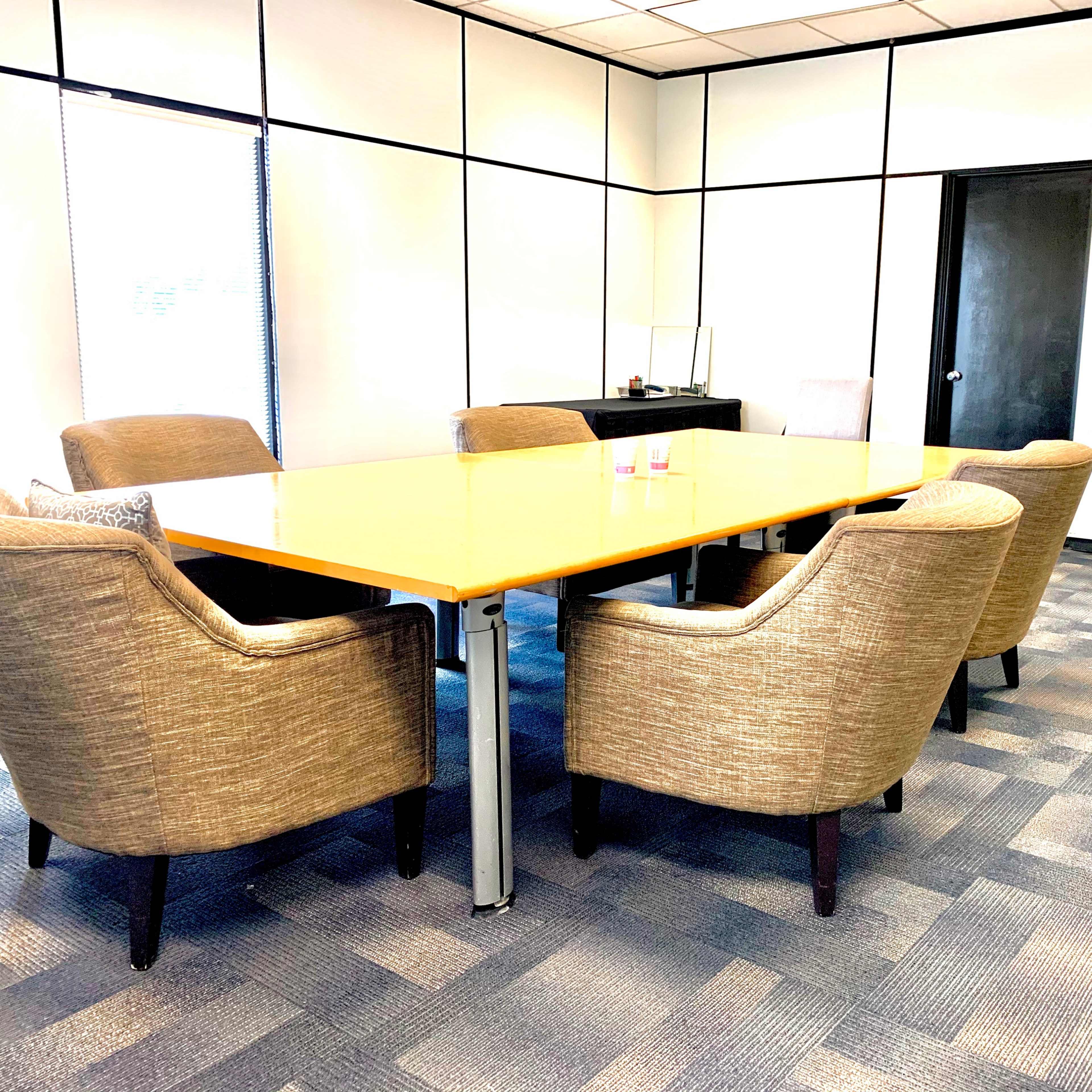 A spacious conference room features a large rectangular table surrounded by six upholstered chairs, with a whiteboard and a plant in the background.