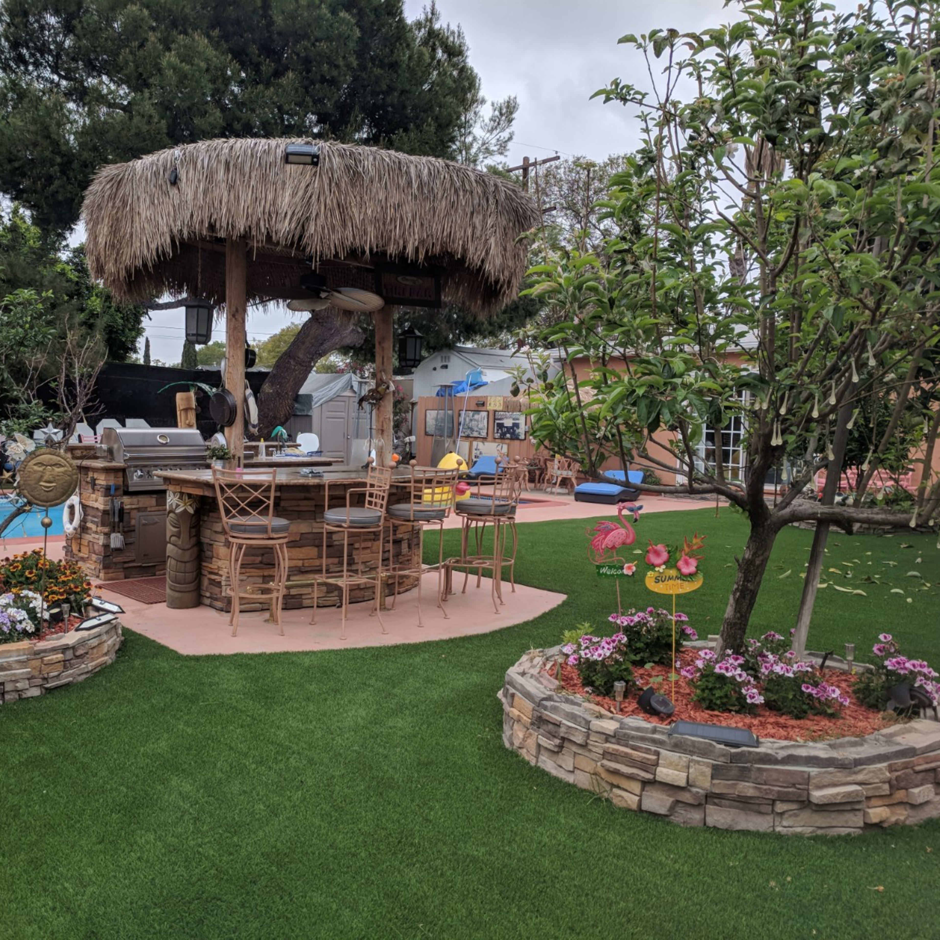 A backyard with a thatched-roof bar surrounded by colorful landscaping and outdoor recreational items.