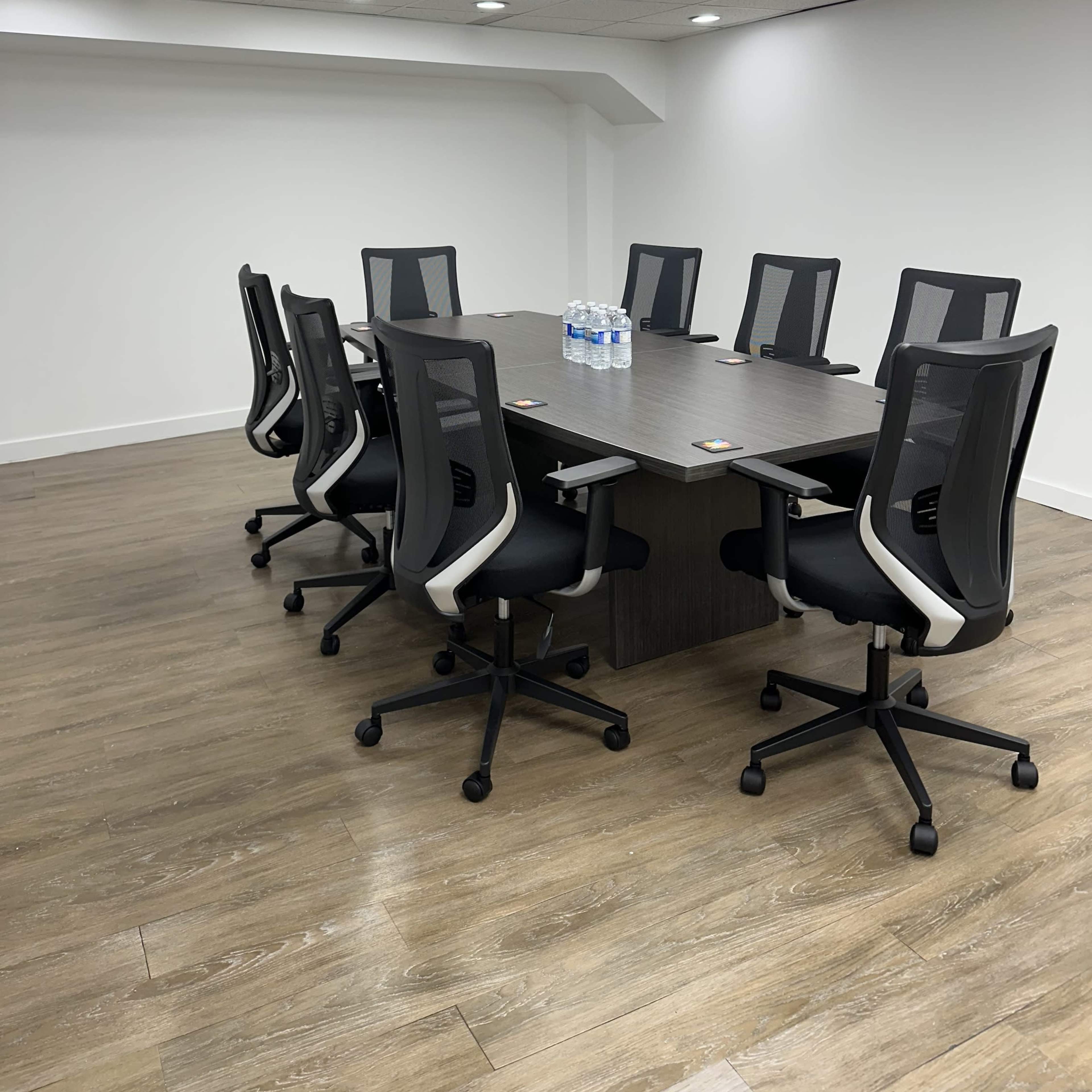 A conference room features a long table surrounded by eight ergonomic chairs and a few bottles of water on the table.