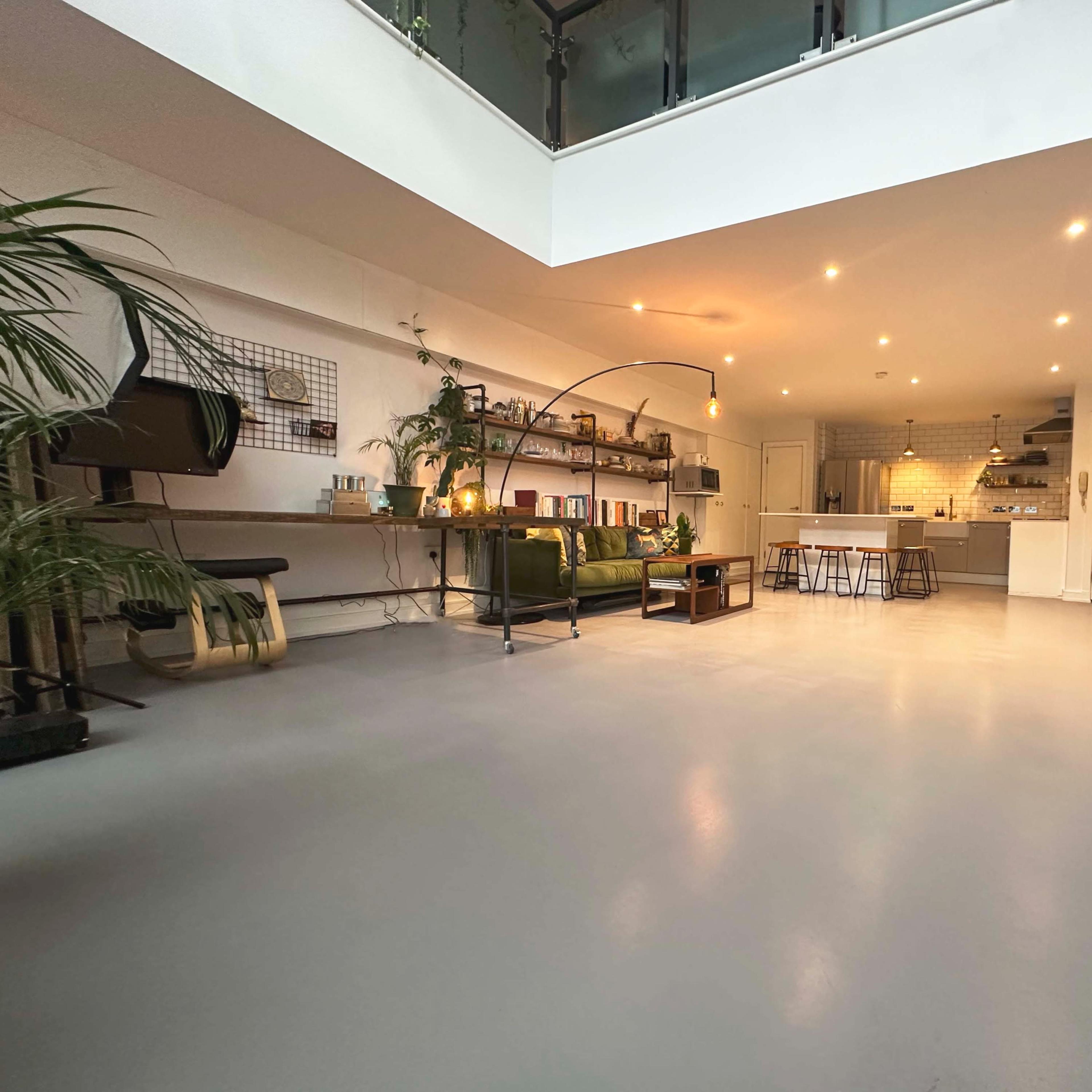 A spacious, modern room featuring a green sofa, a wooden table with stools, and a well-lit kitchen area in the background.