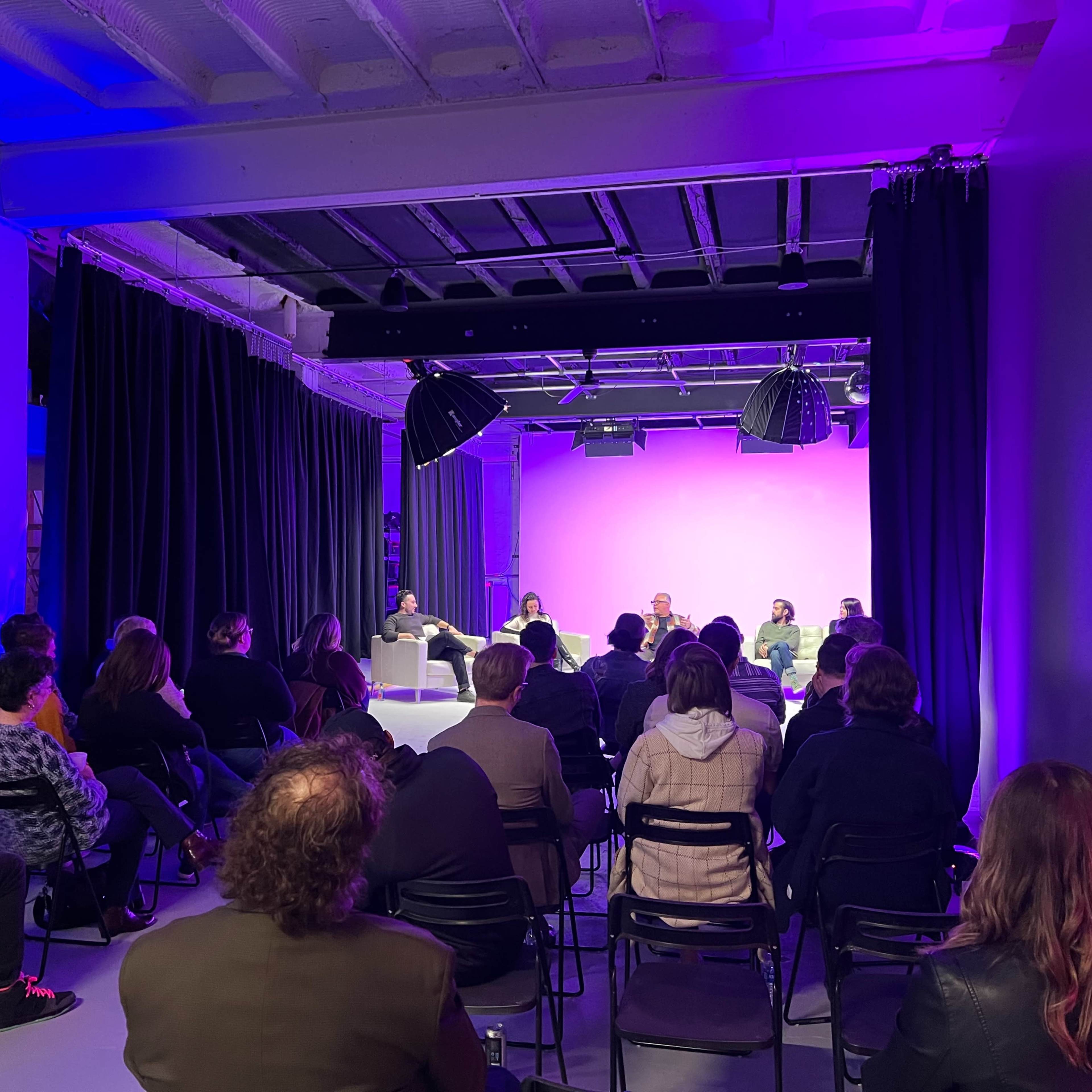 A panel discussion is taking place in a modern venue, with seated audience members facing a group of speakers on a stage illuminated by purple lighting.