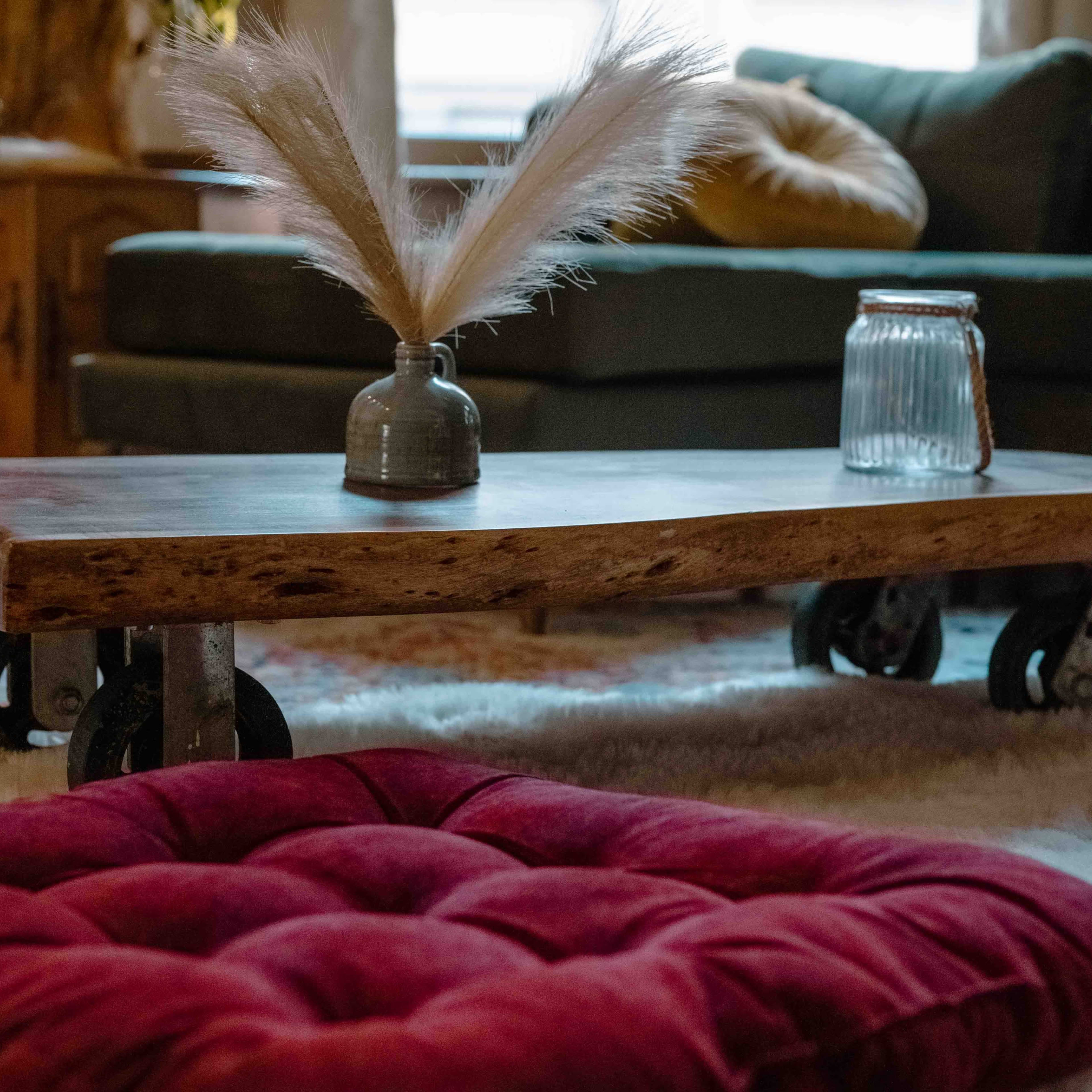 A wooden coffee table on wheels is positioned in a living room, with a small vase and a glass jar atop it, surrounded by plush, red cushions on a plush rug.