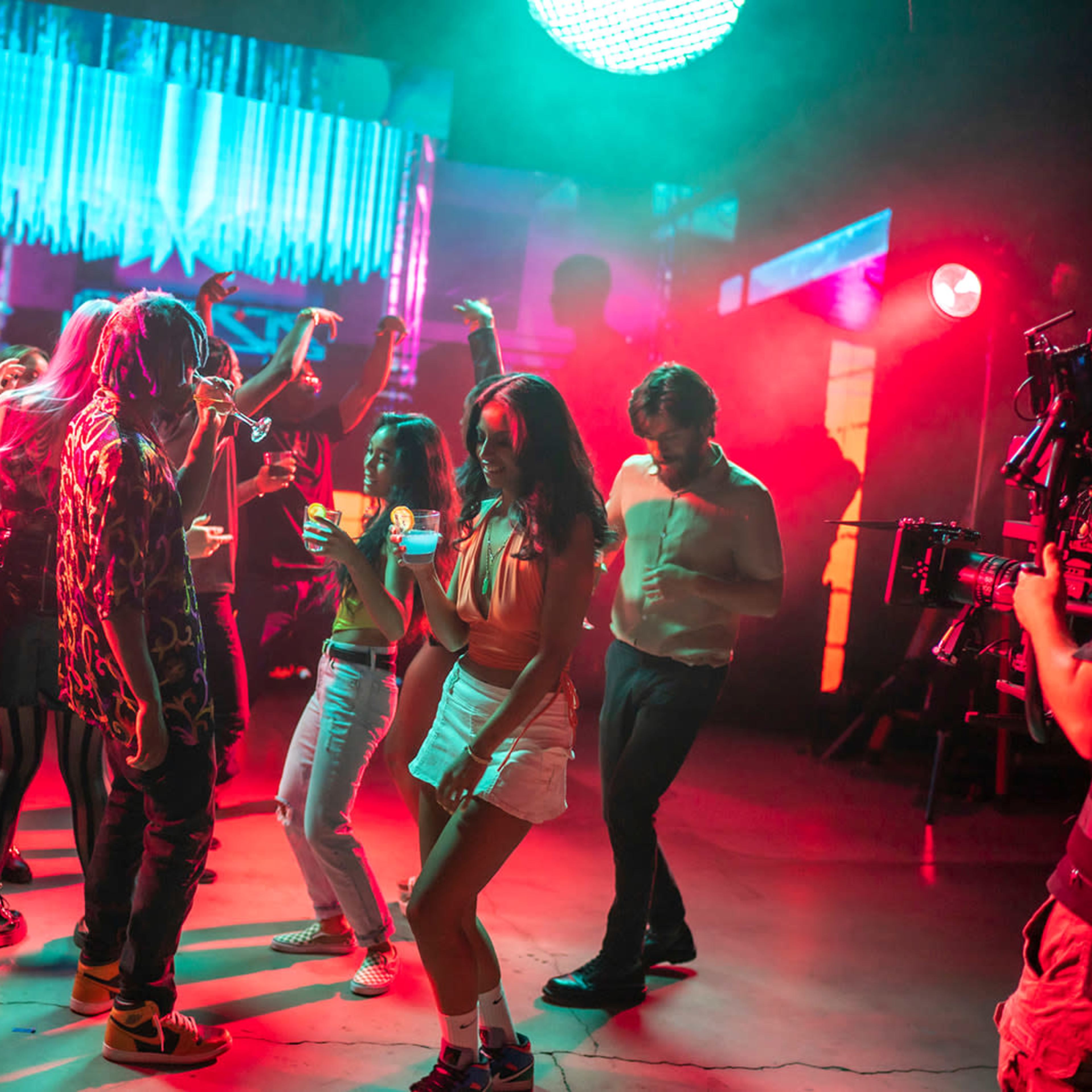 A group of people is dancing under colorful lights in a nightclub setting while a camera crew captures the scene.