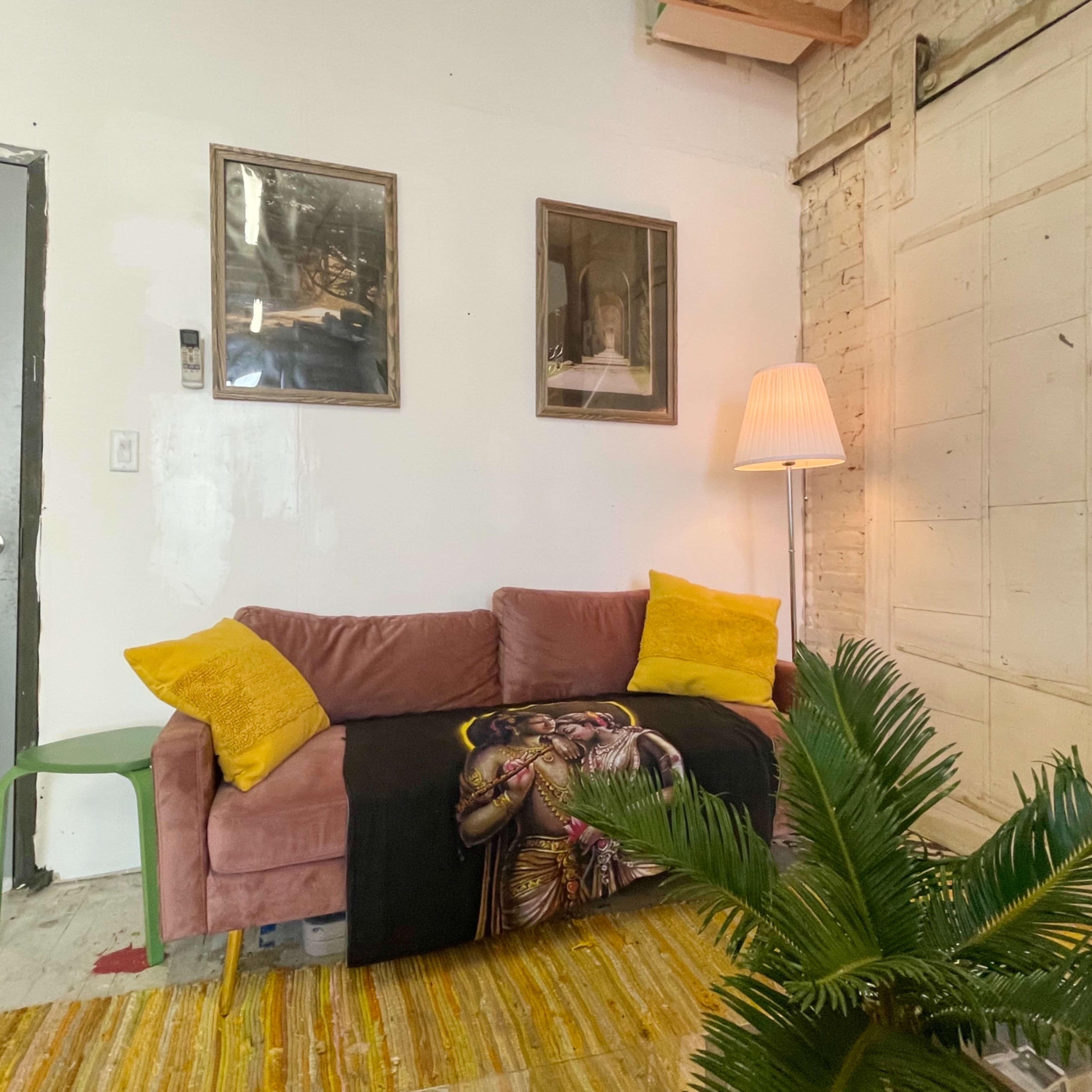 A cozy living space with a pink sofa adorned with yellow pillows, a patterned throw, a standing lamp, two framed pictures on the wall, and a green chair next to a potted plant.