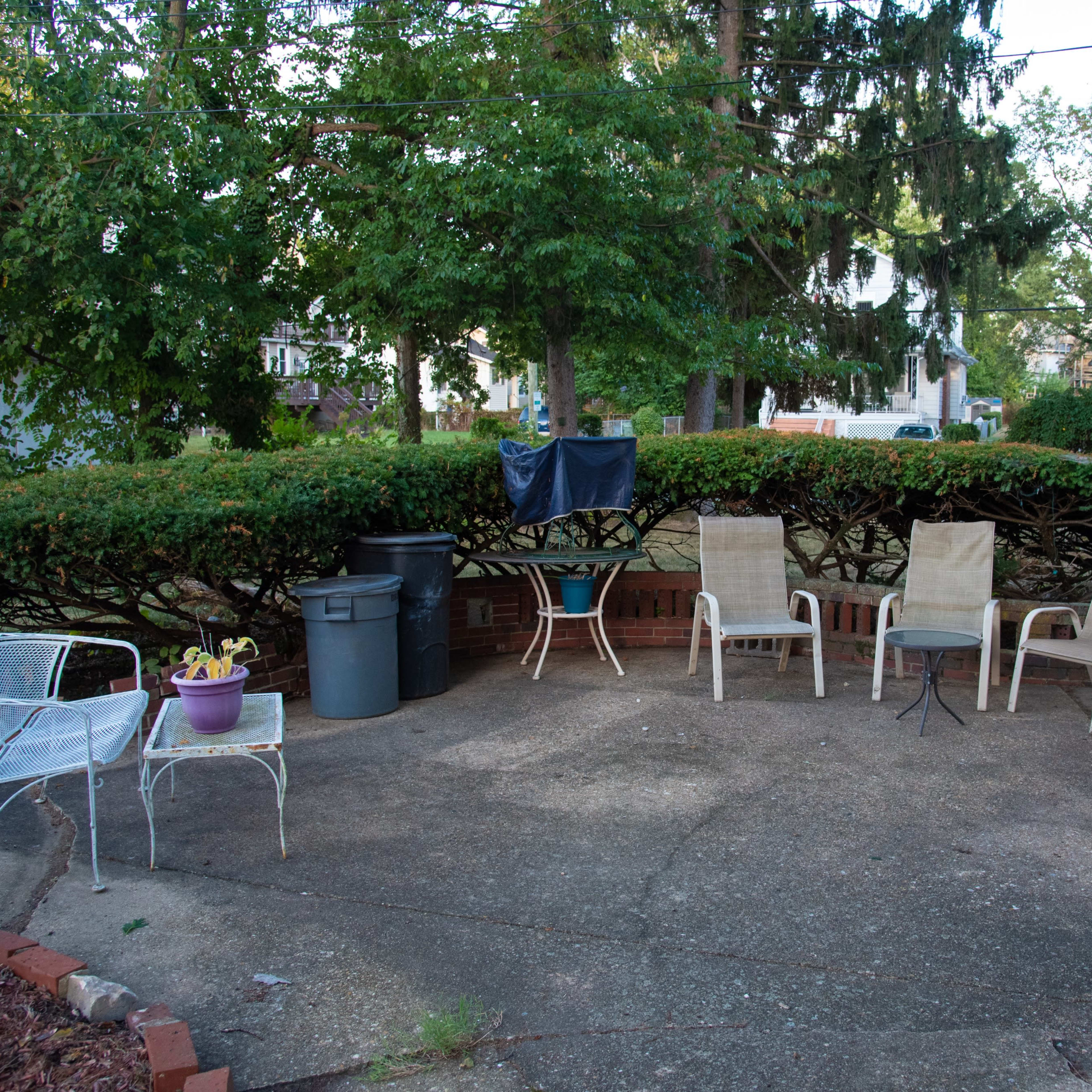 A concrete patio is surrounded by bushes and features several chairs, a trash bin, and potted plants.