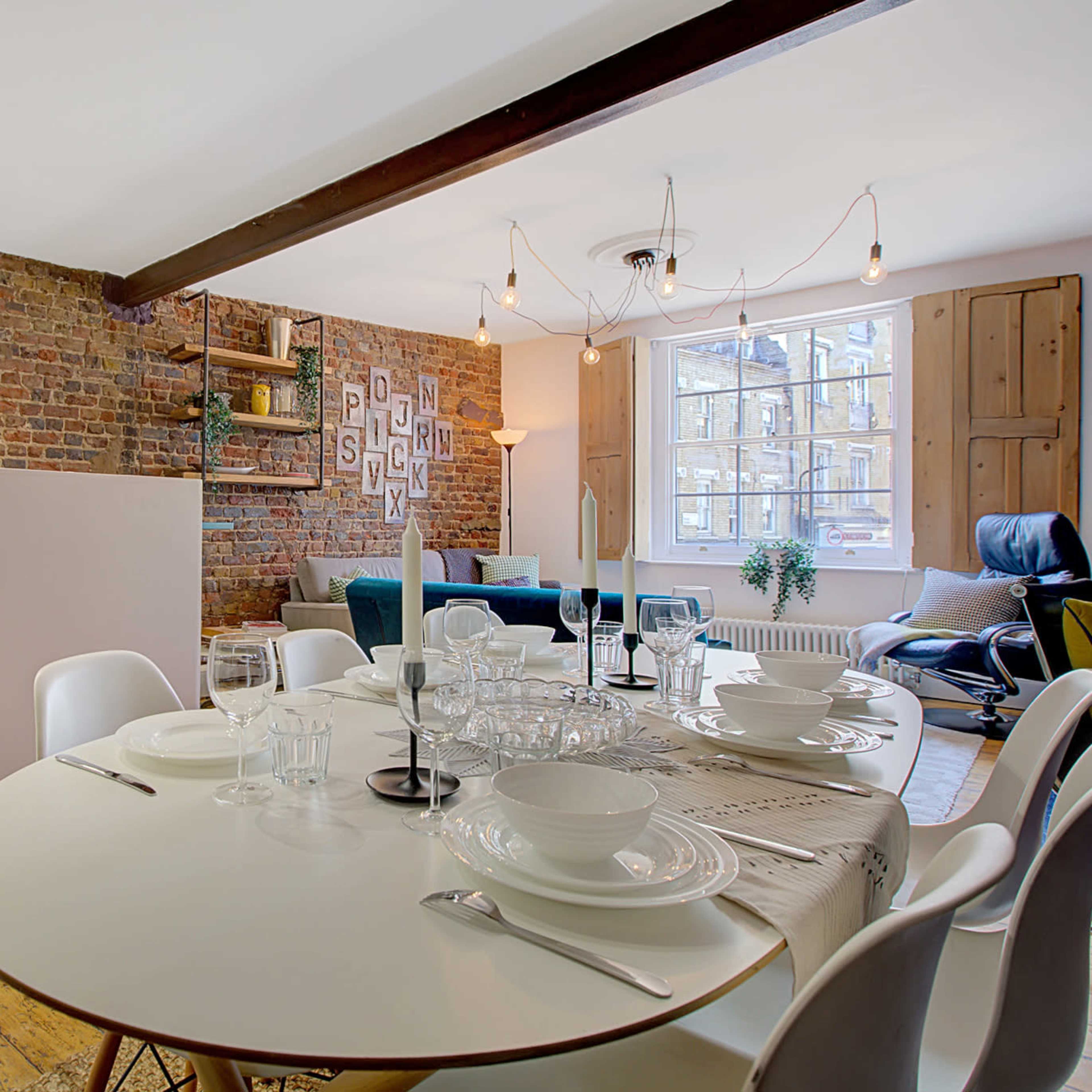 A modern kitchen and dining area features a round table set for a meal, surrounded by white chairs and a backdrop of exposed brick walls.