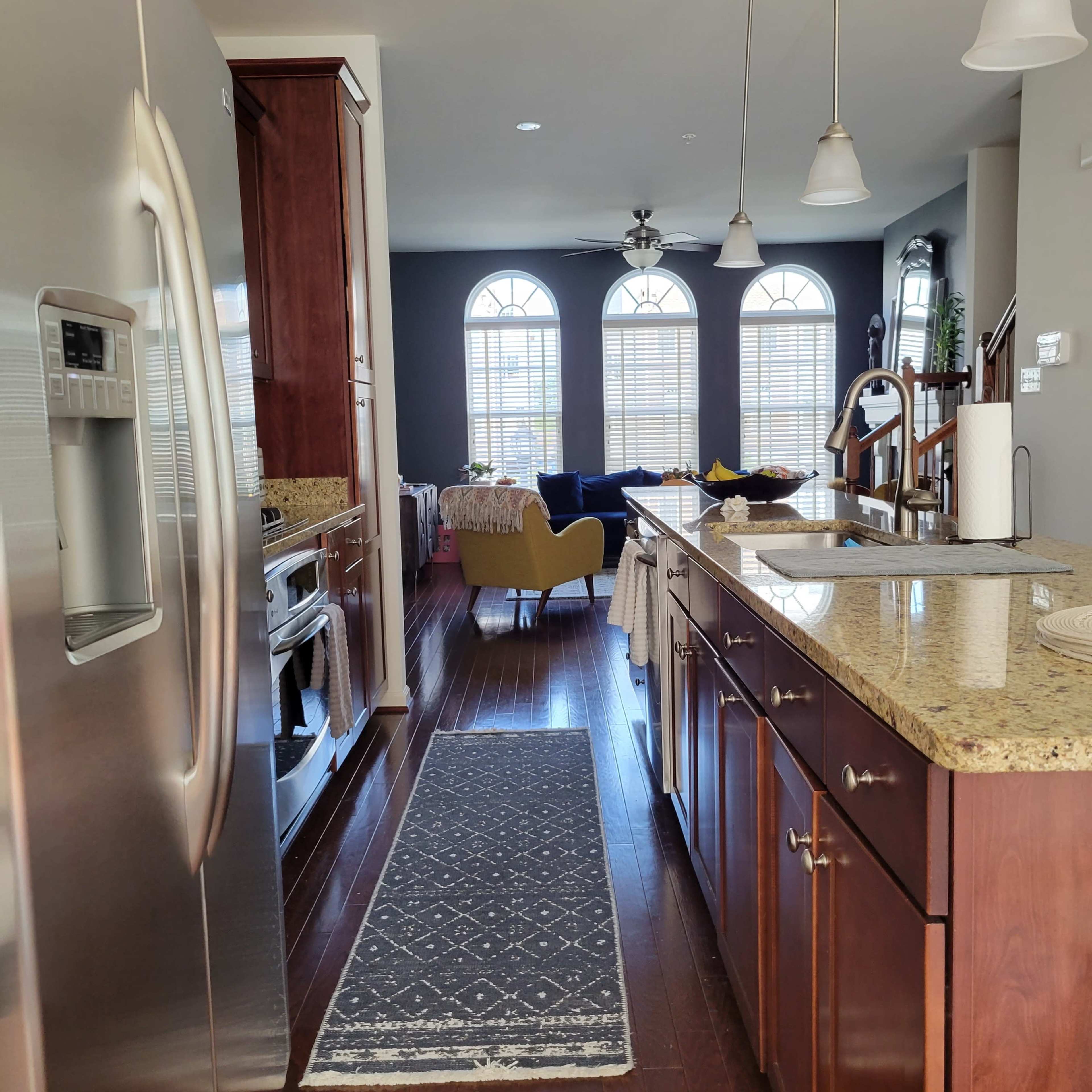 A kitchen with dark wood cabinetry, a stainless steel refrigerator, and a patterned rug running along the floor, leading to a living area with large windows and a sofa in the background.