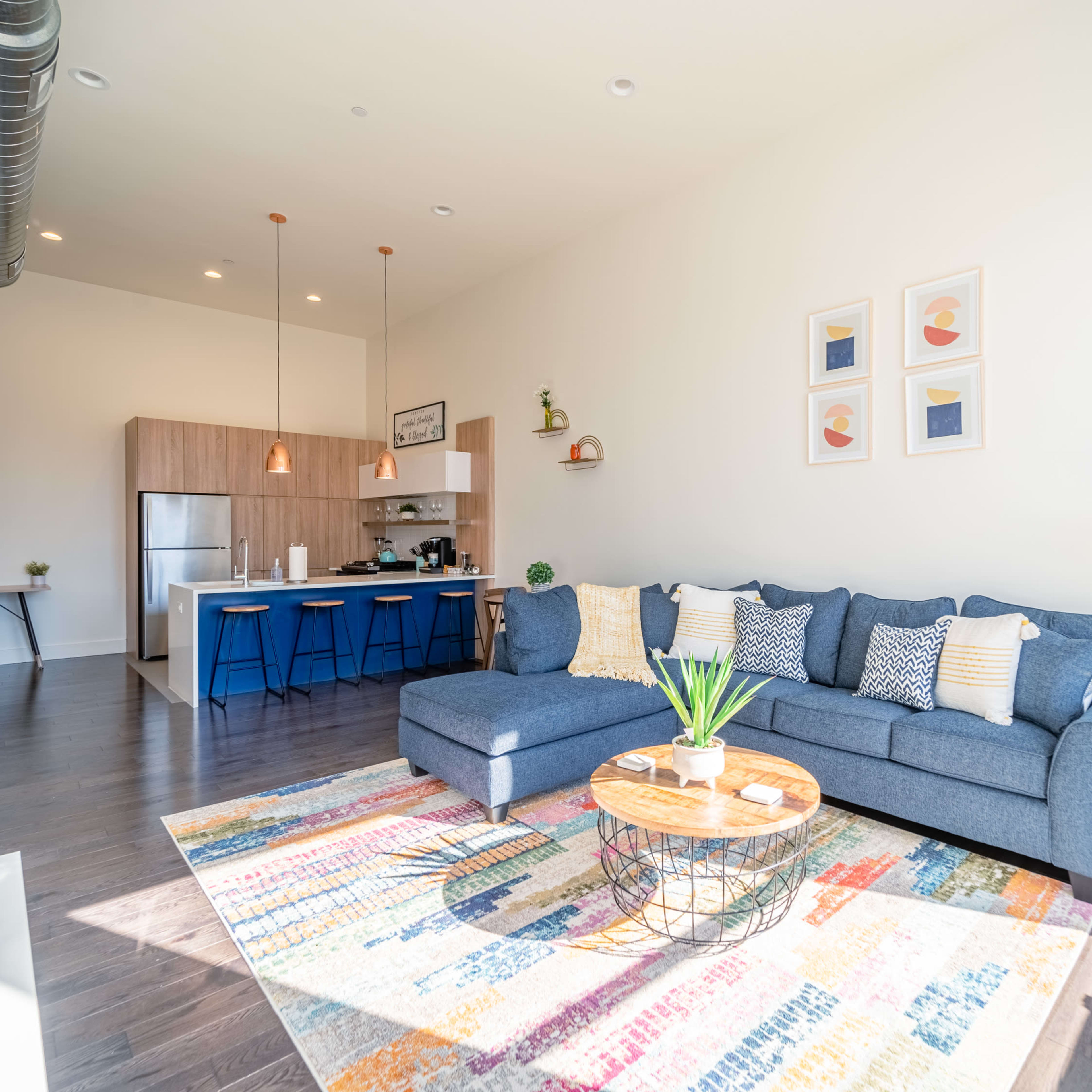 A modern living room features a blue sectional sofa, a round coffee table with plants, and a view of an open kitchen with wooden cabinets and bar seating.