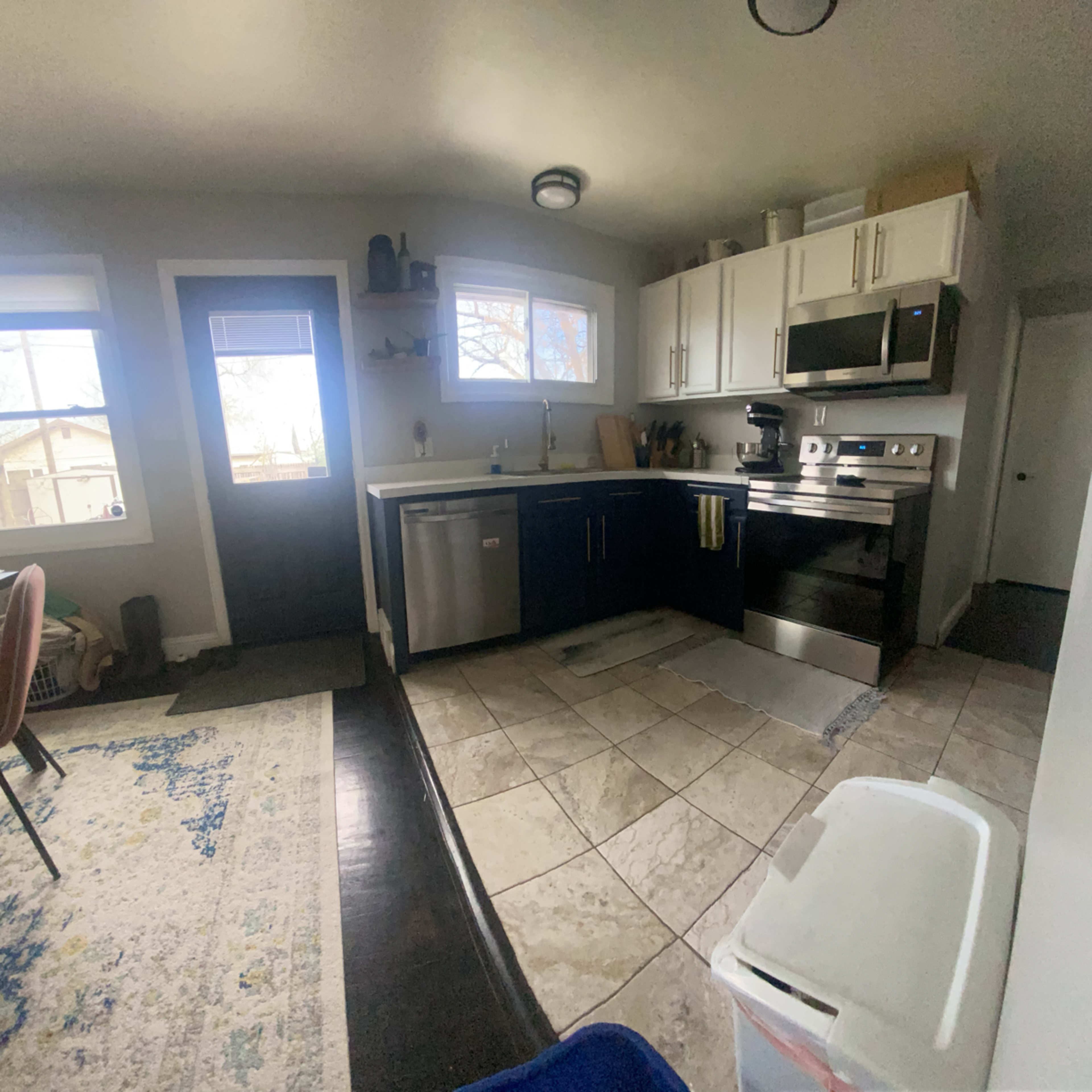 A modern kitchen with stainless steel appliances, a dark countertop, and tile flooring, connected to a dining area with a rug.