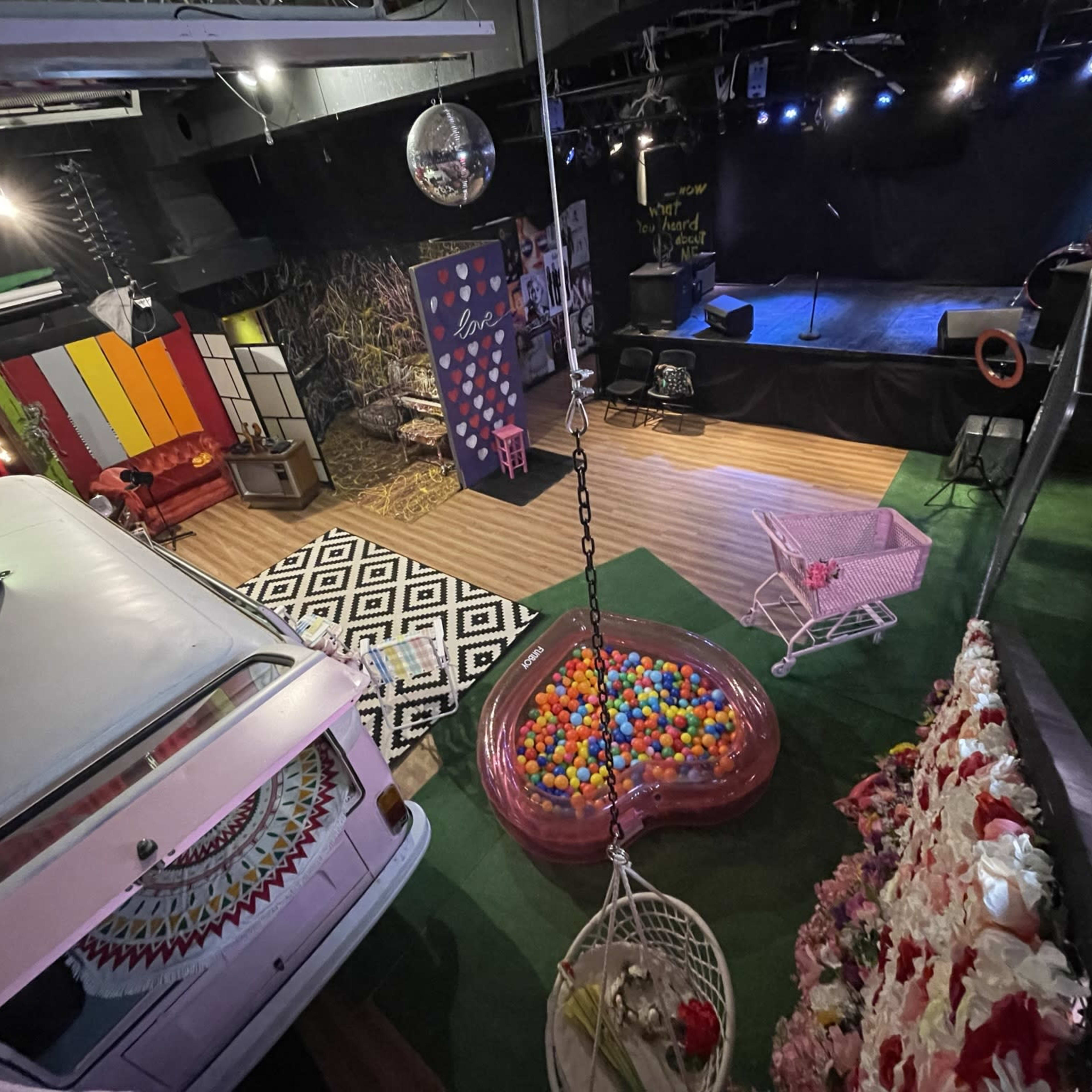 A colorful performance space featuring a retro van, a ball pit, hanging chairs, and a stage with blue lighting in the background.