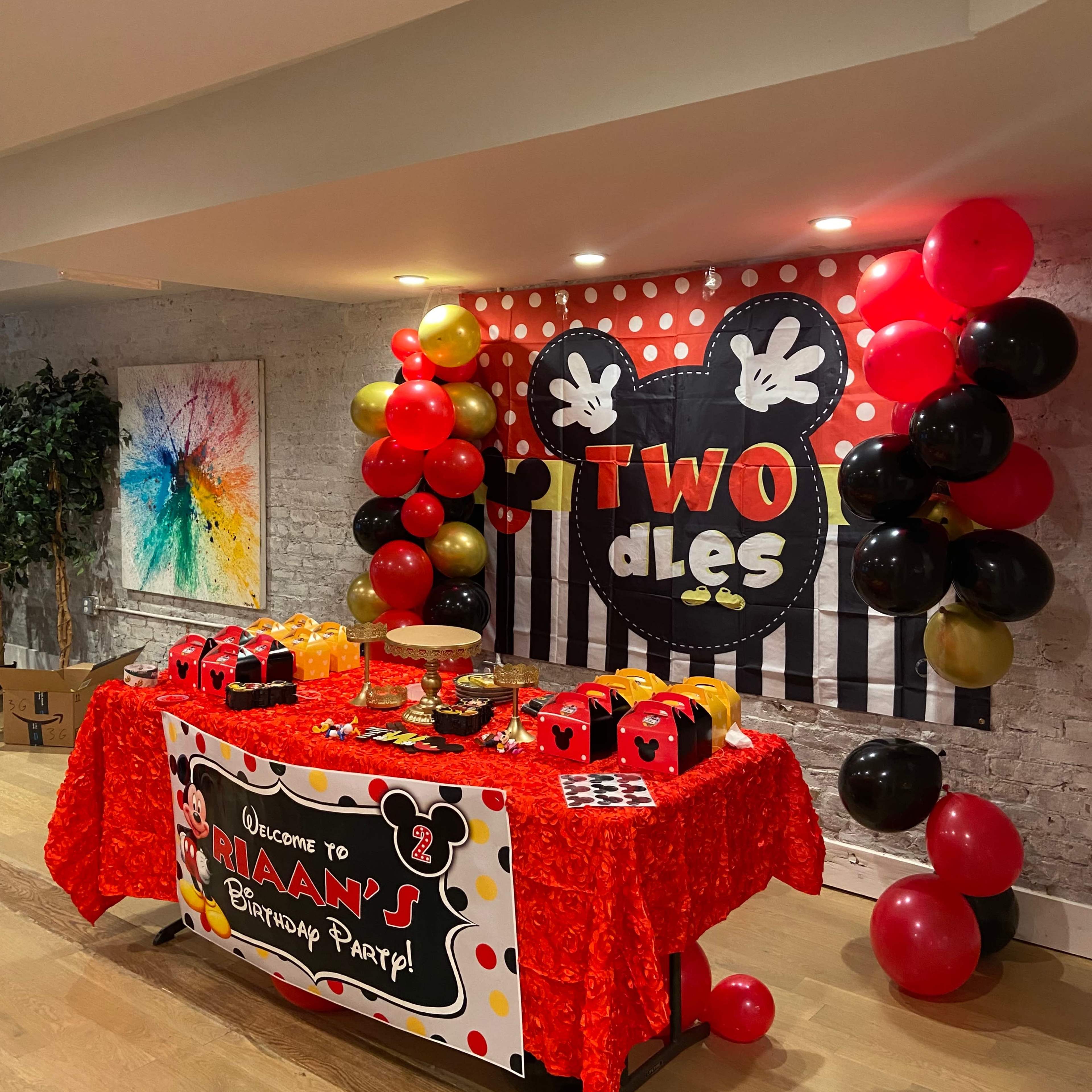 A birthday party setup features a table decorated with a red tablecloth, themed cakes, and balloons, alongside a backdrop with a large Minnie Mouse design and festive banners.