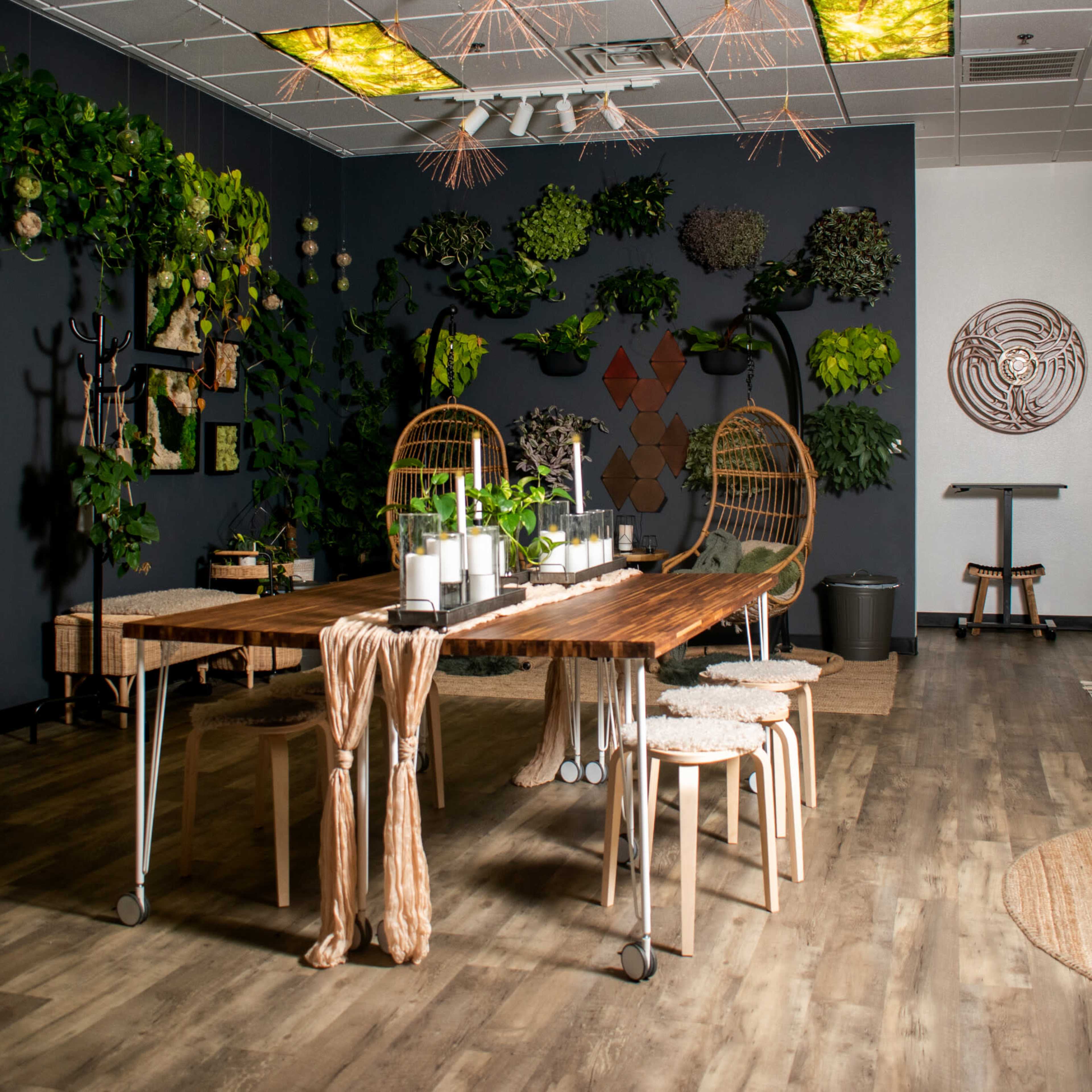 A brightly lit indoor space features a long wooden table surrounded by stools, with a backdrop of greenery and decorative wall art.